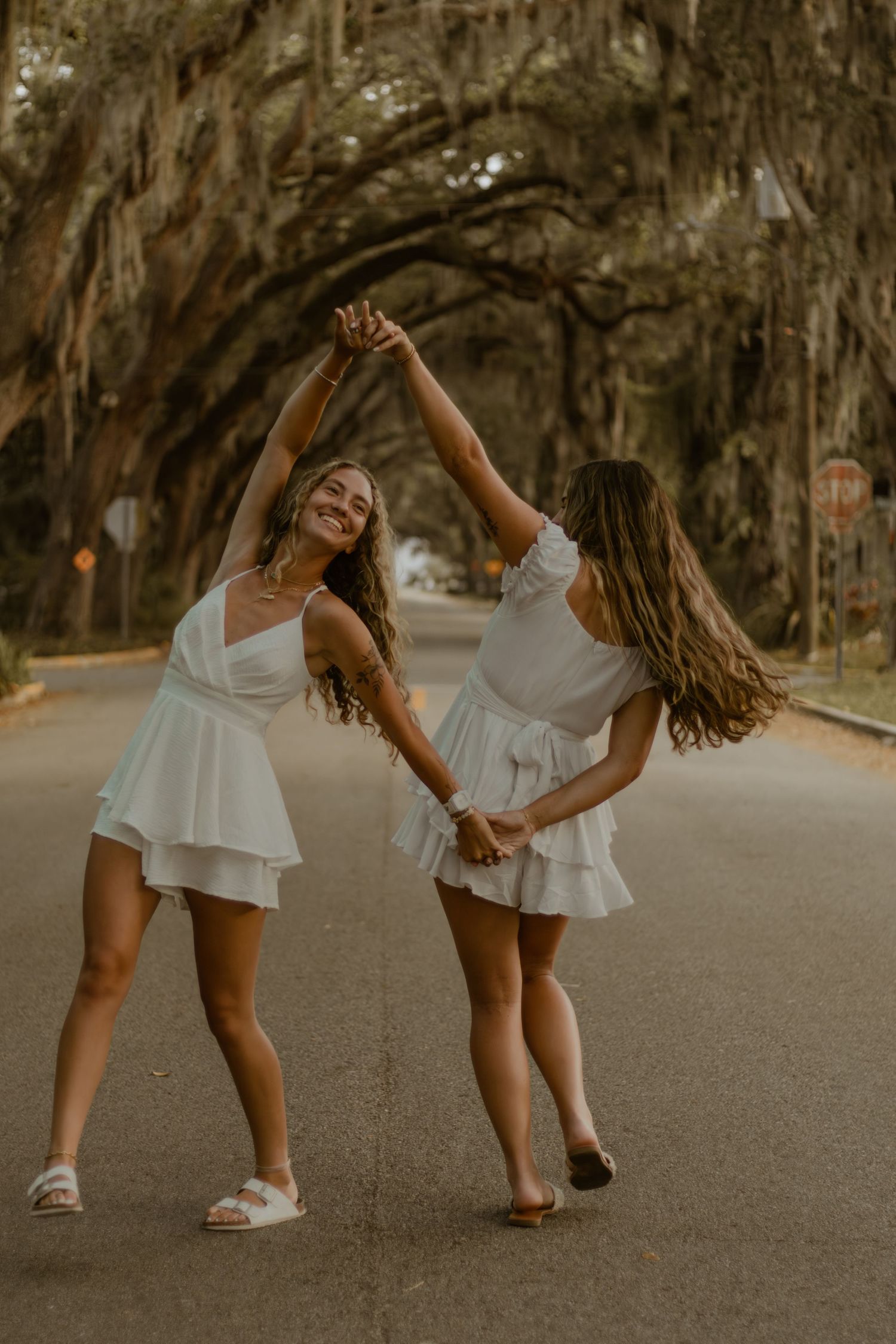Two friends in white dresses dancing and twirling on a tree-lined street.