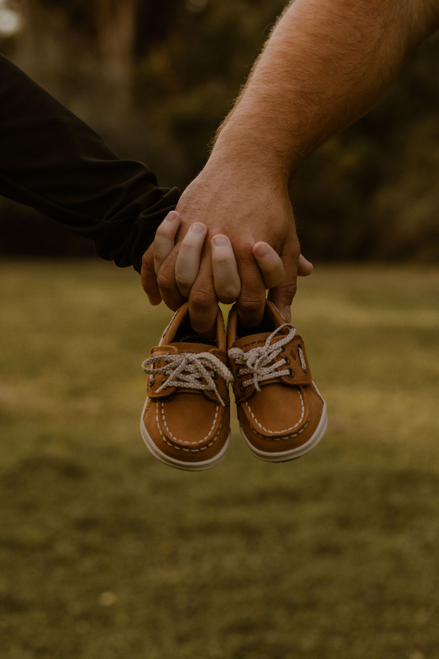 Hands holding small brown leather baby moccasins against grass.