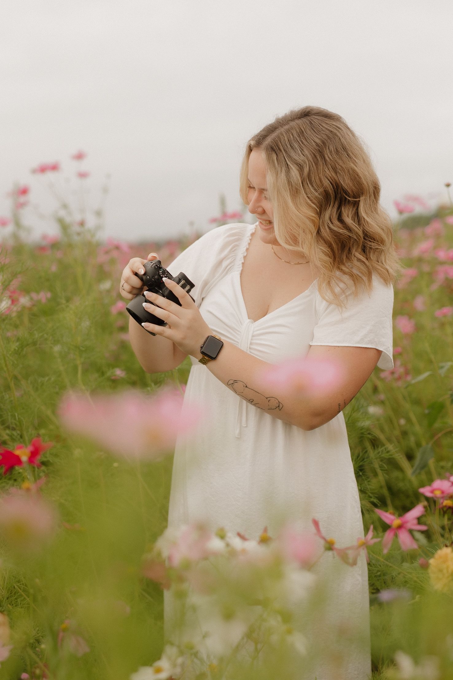 A photographer checks their camera while standing in a field of pink wildflowers on a sunny day.