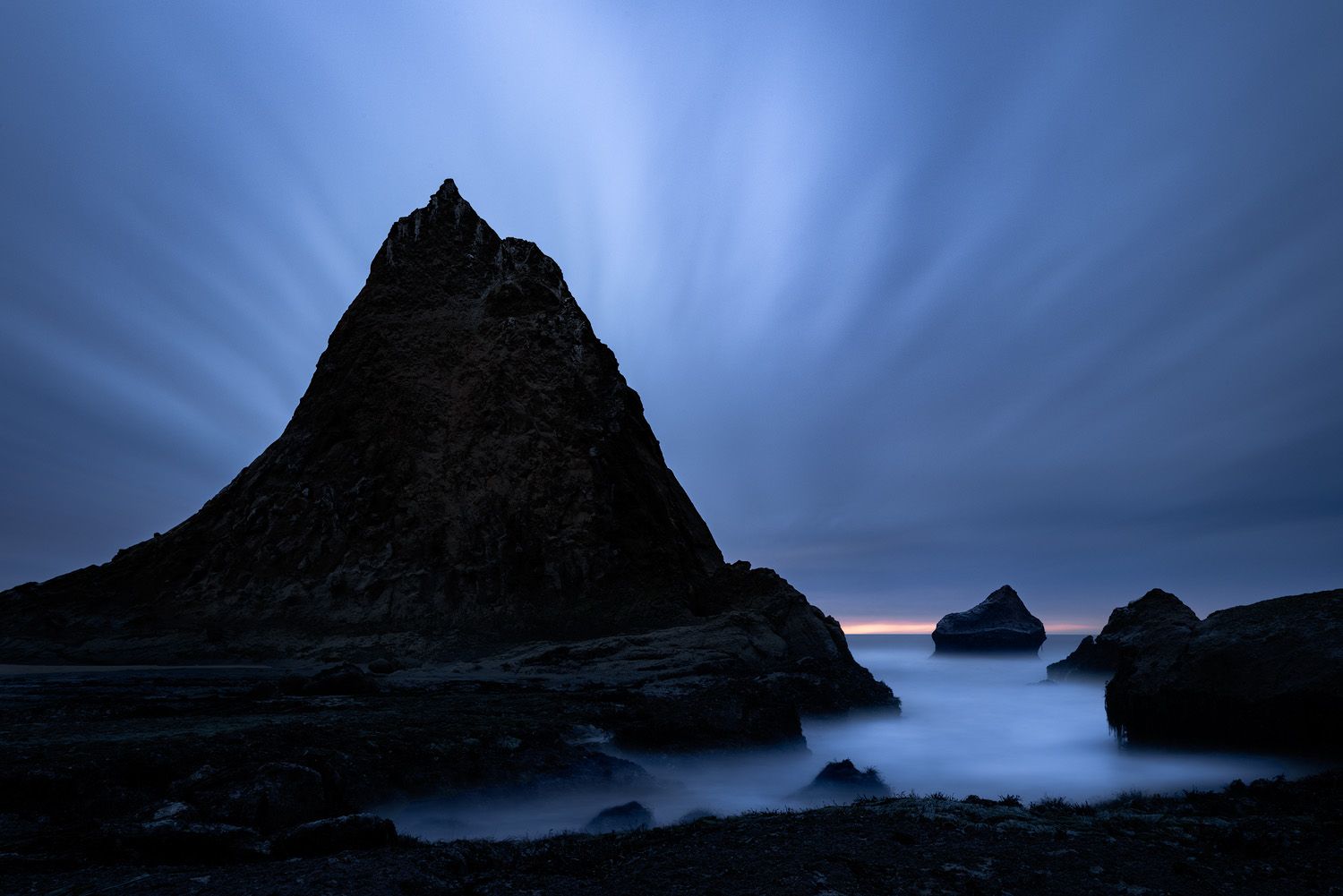 Dramatic silhouette of a pyramid-shaped coastal rock formation against a moody blue twilight sky.