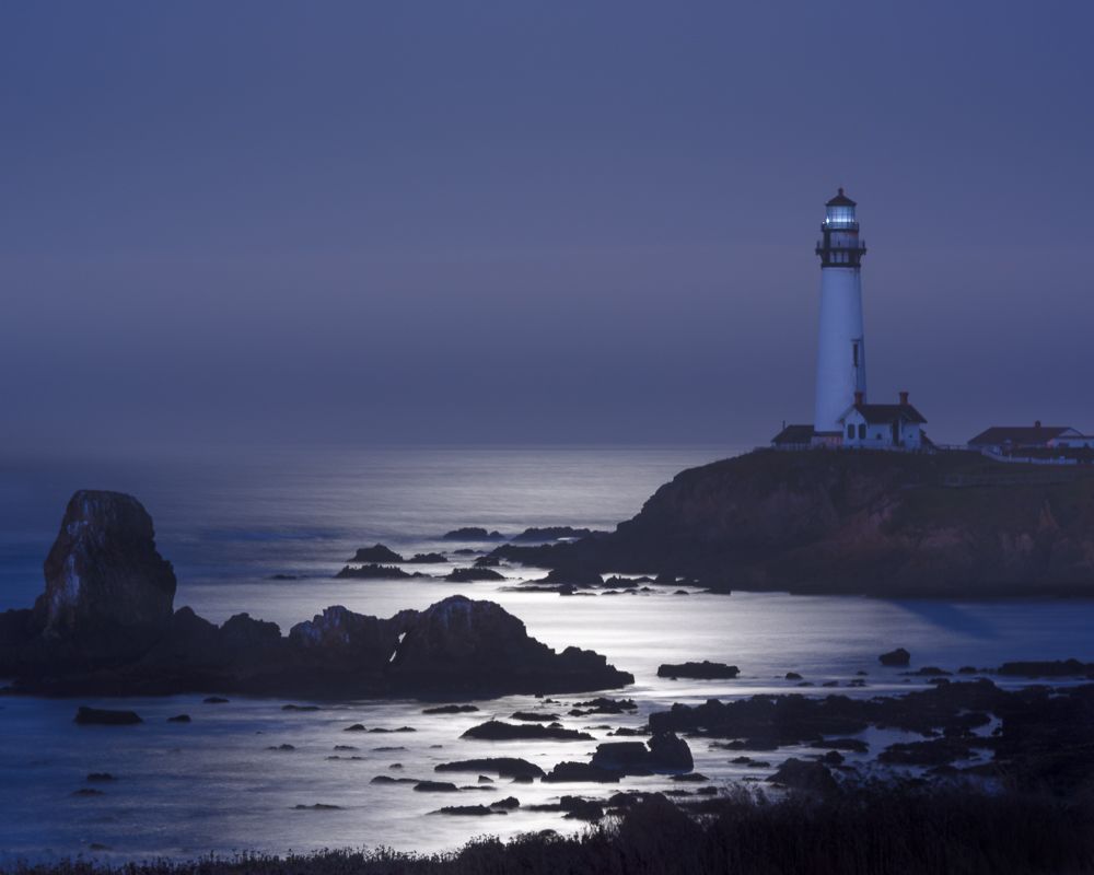 Moonlit coastal landscape with lighthouse and rocky shoreline silhouetted against dark waters.