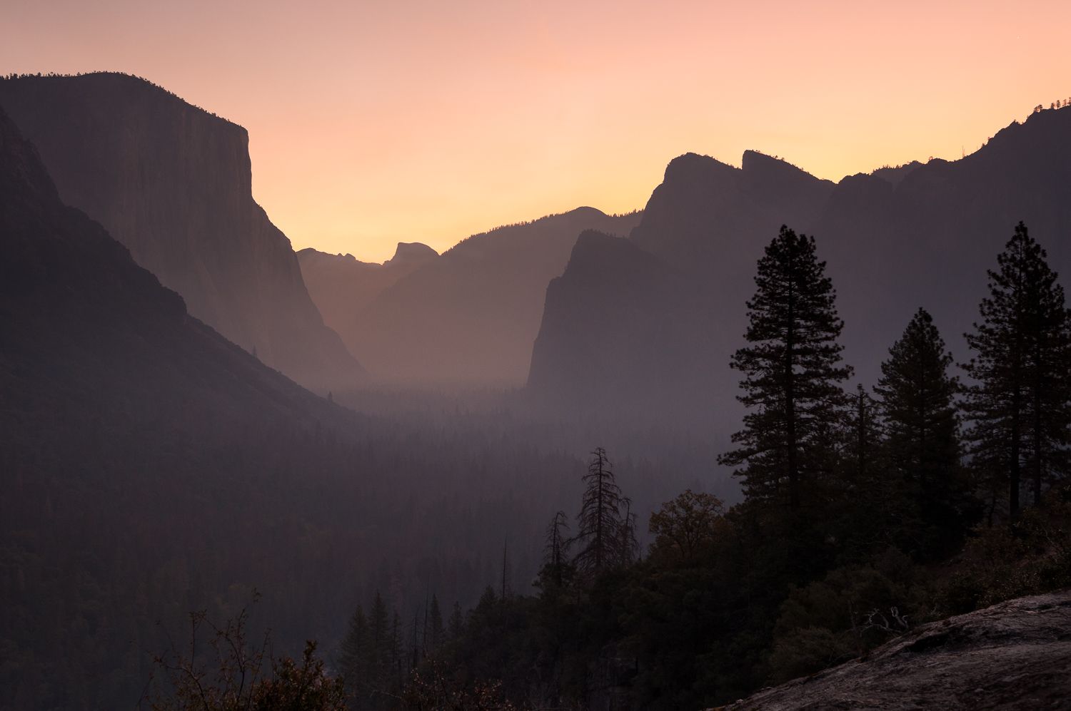 Sunrise silhouettes mountain peaks in Yosemite Valley with pink and purple hues.
