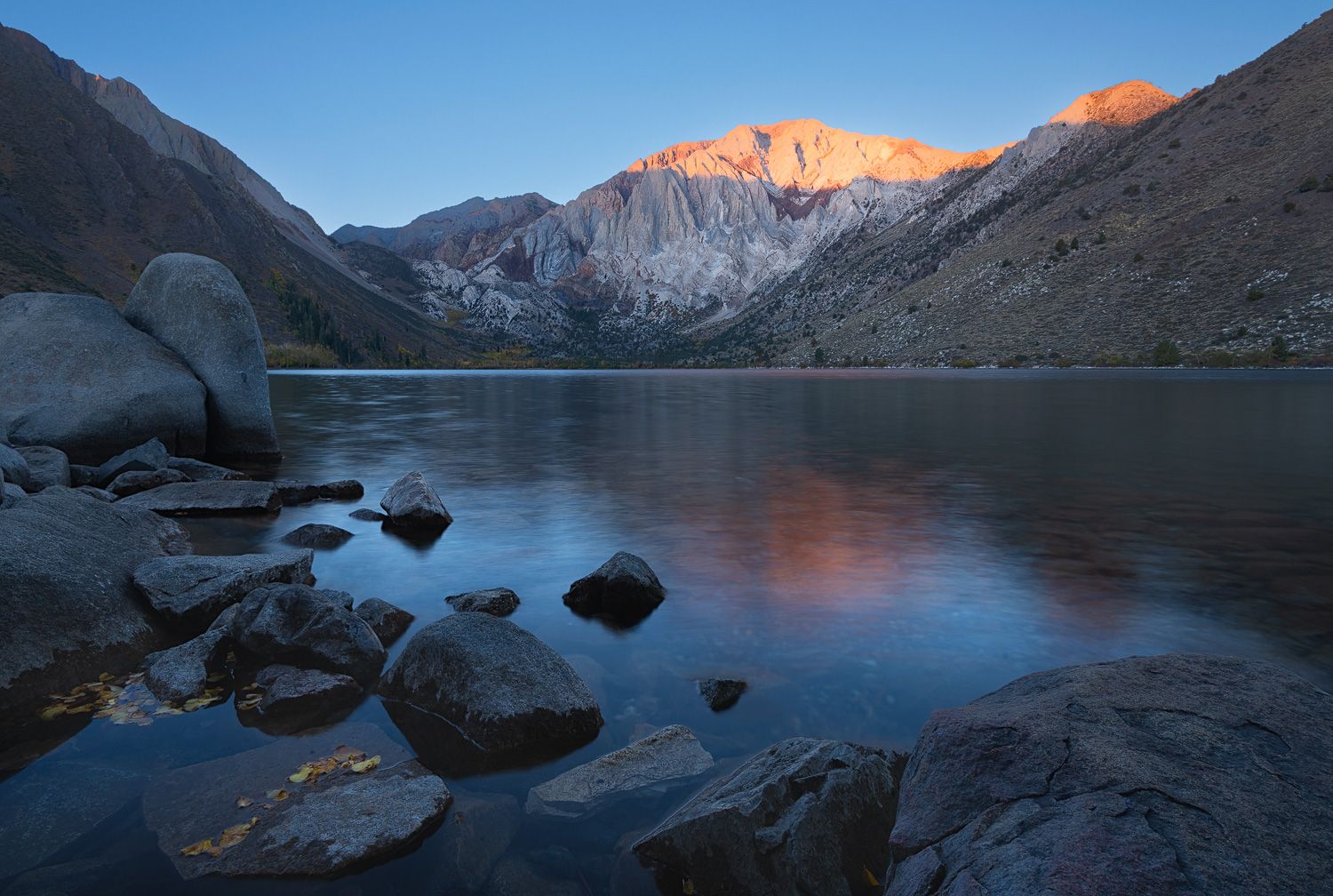 Mountain lake reflects dramatic sunrise light on surrounding peaks and rocky shoreline.