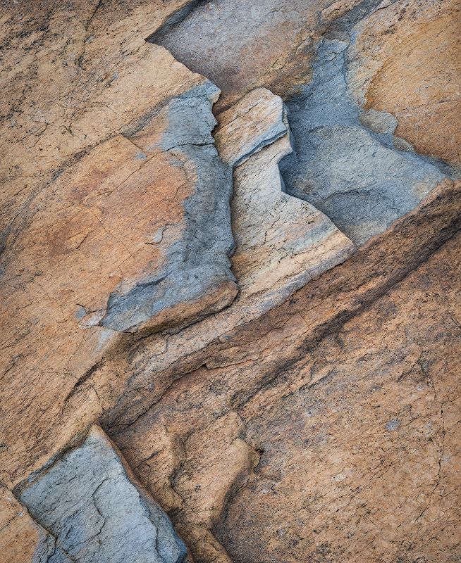 Abstract pattern of layered sandstone showing tan and bluish-gray geometric shapes and cracks.