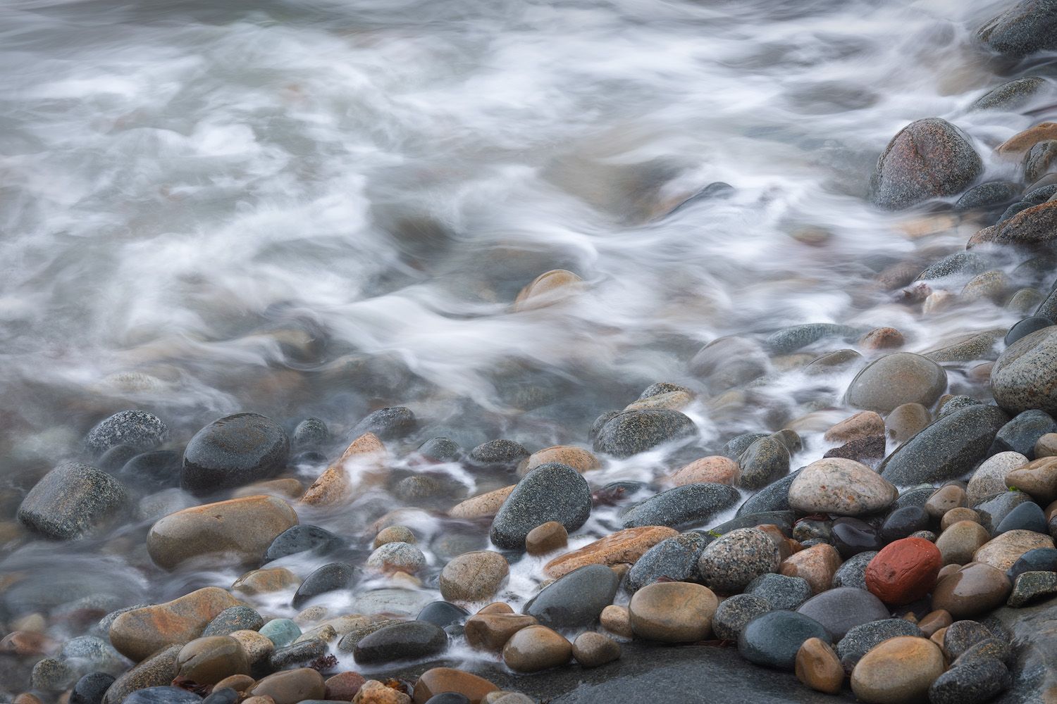 Long exposure of smooth waves washing over colorful rounded stones on a rocky beach.
