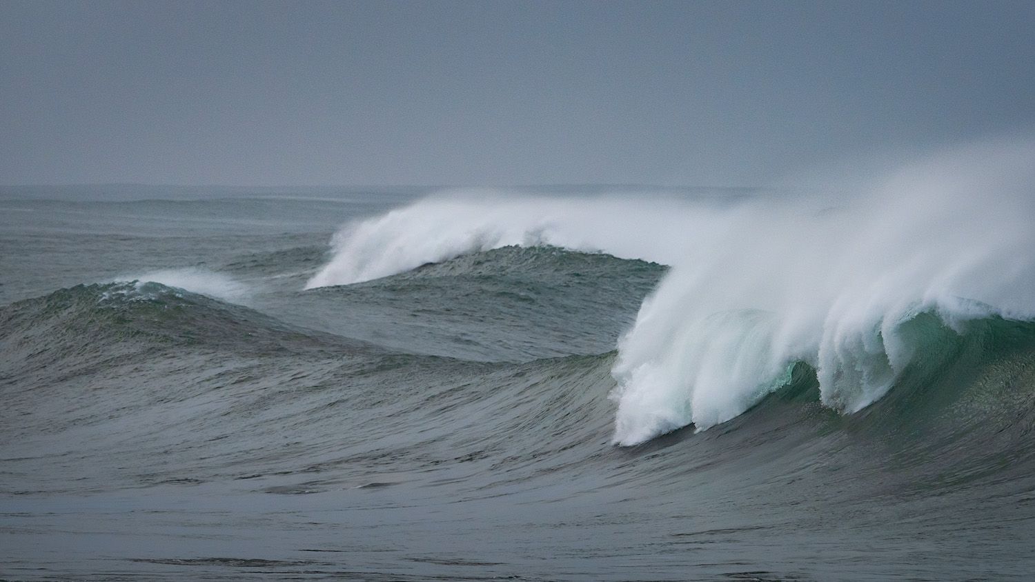 Series of powerful ocean waves breaking with white spray against stormy gray skies.