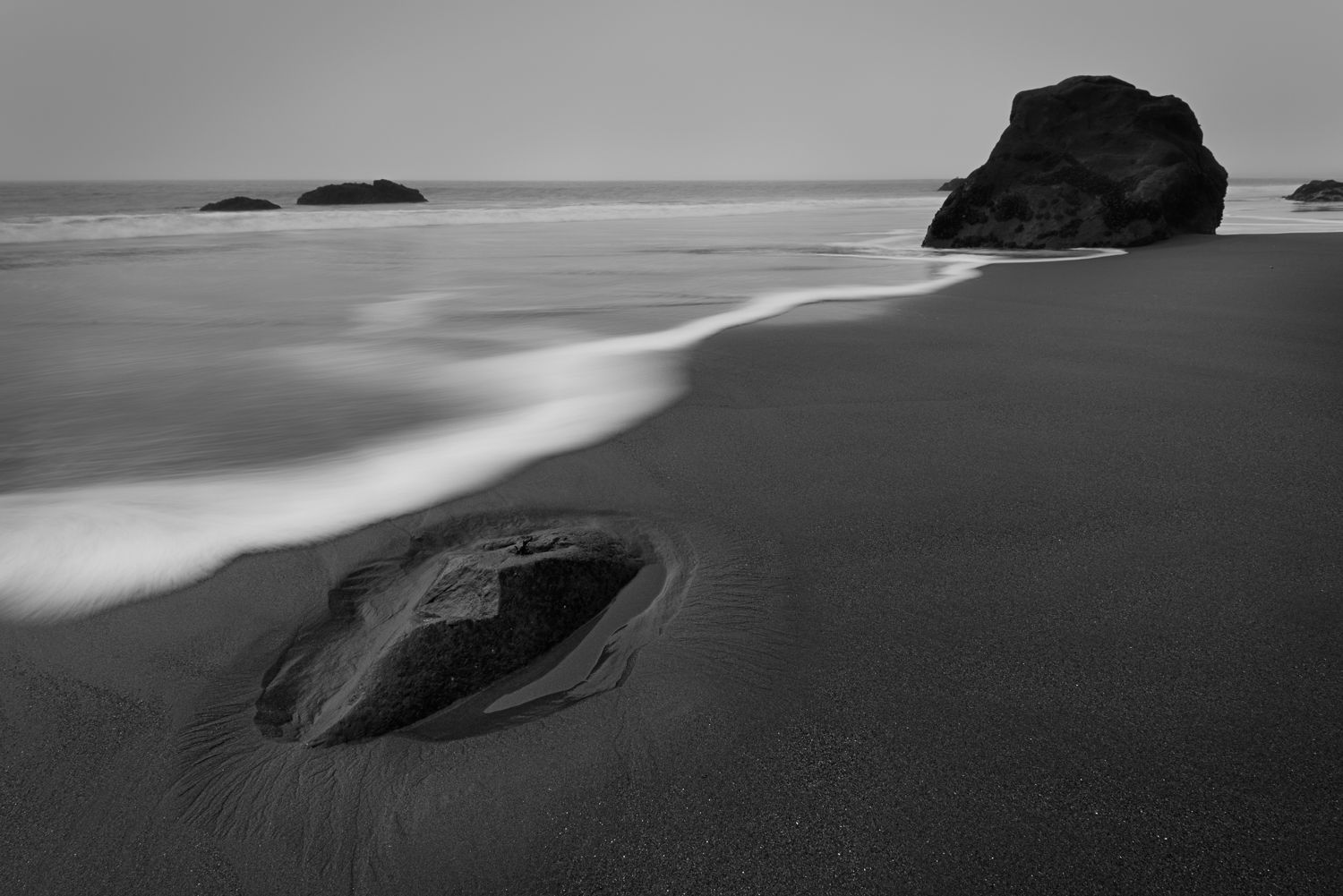Dramatic black and white seascape with dark rocks and white surf along a misty shoreline.