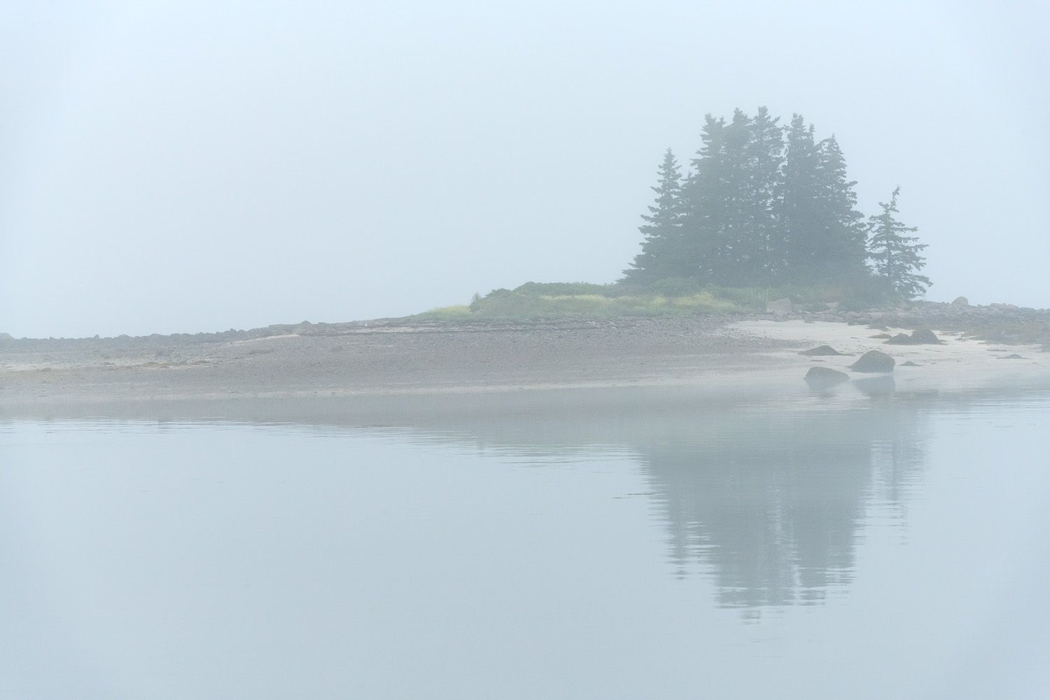 Misty fog surrounds a small forested island on a tranquil lake during a calm morning.