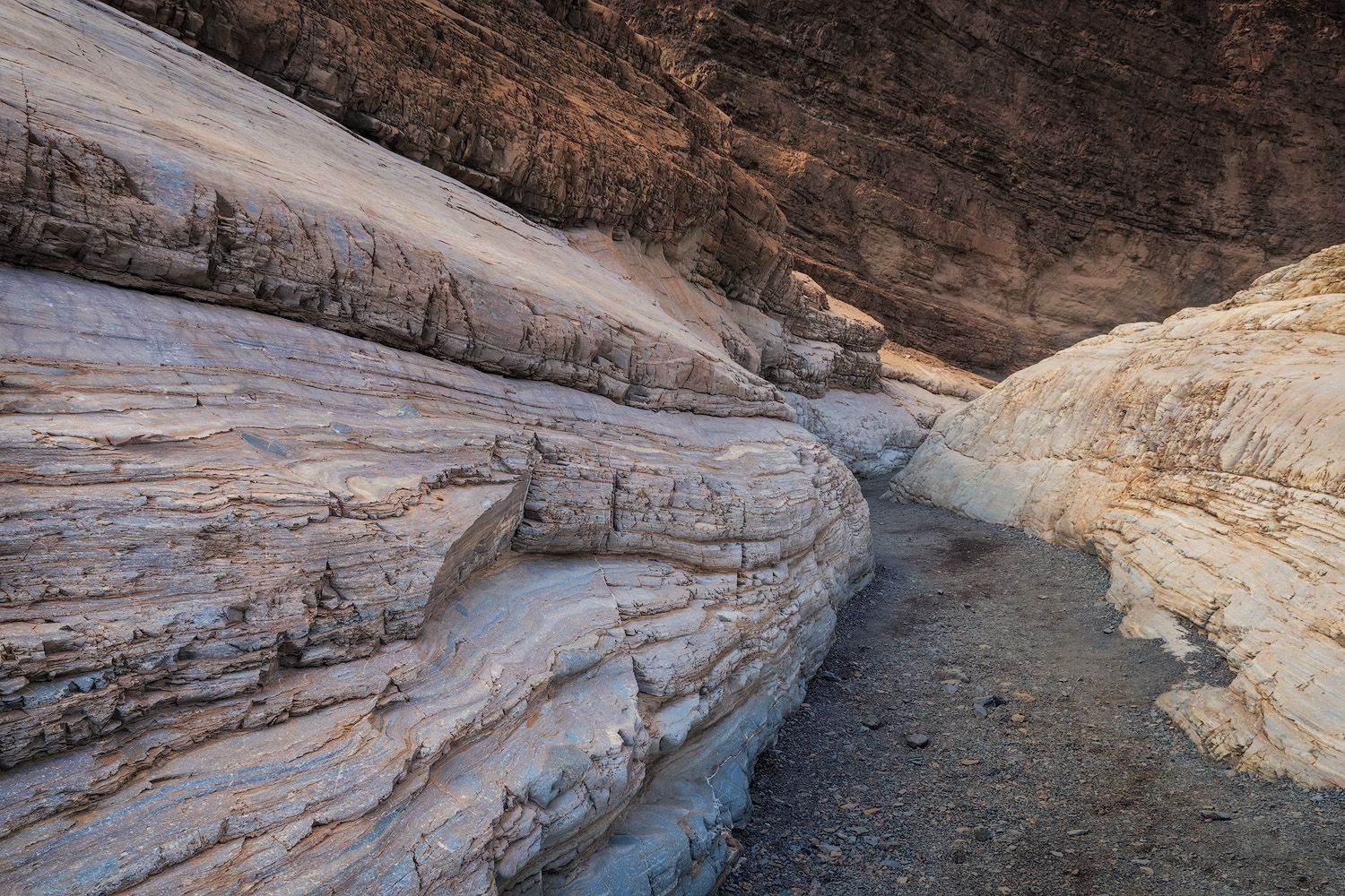 Layered white rock formations in a narrow desert canyon with sandy floor and warm golden tones.