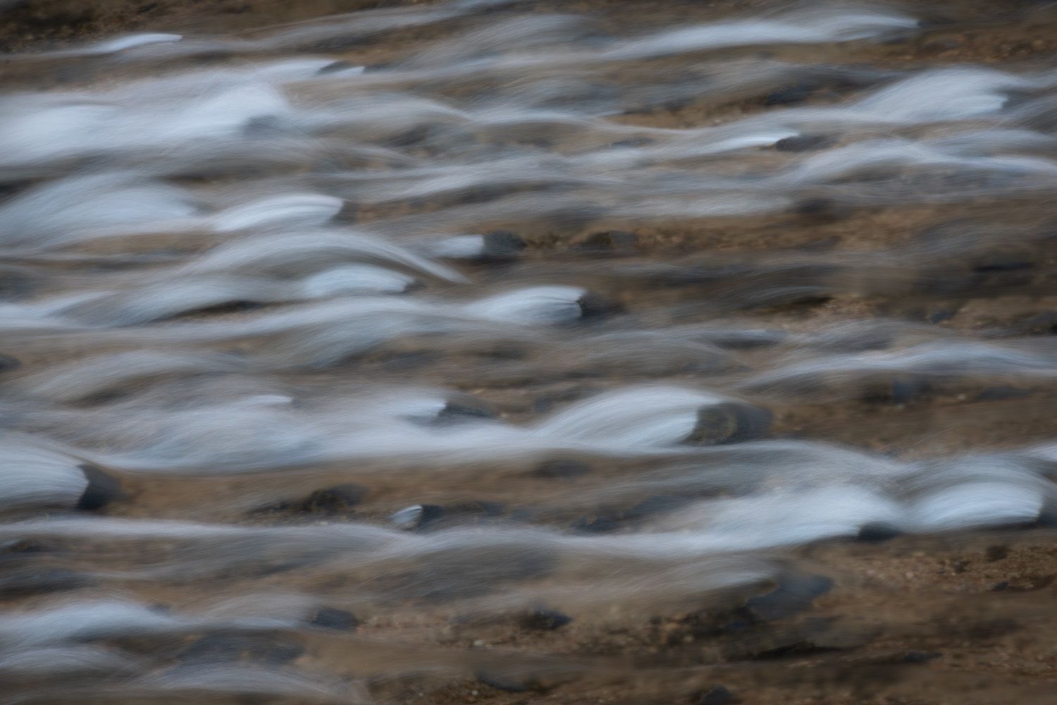 Long exposure photograph of flowing water creating smooth white streaks over brown rocks in a stream.
