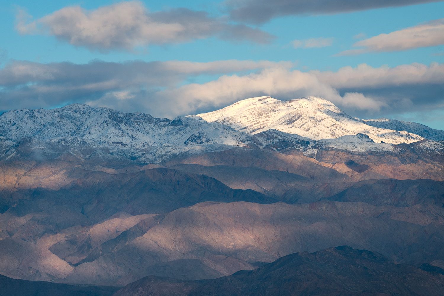 Snow-capped mountain peak rising above layered brown hills under a partly cloudy blue sky in a dramatic landscape.