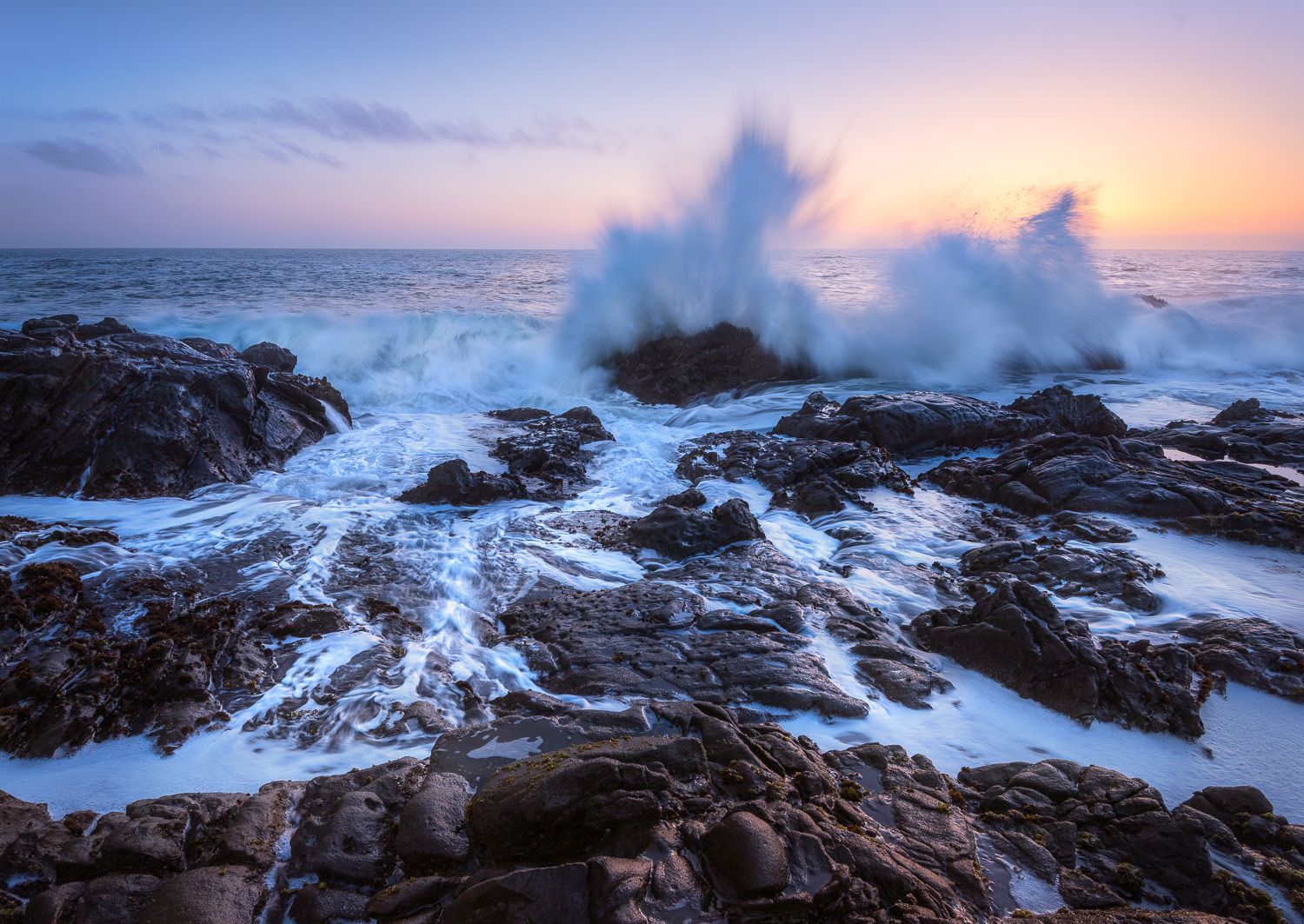 Powerful ocean waves crash against rocky coast during a vibrant sunset.