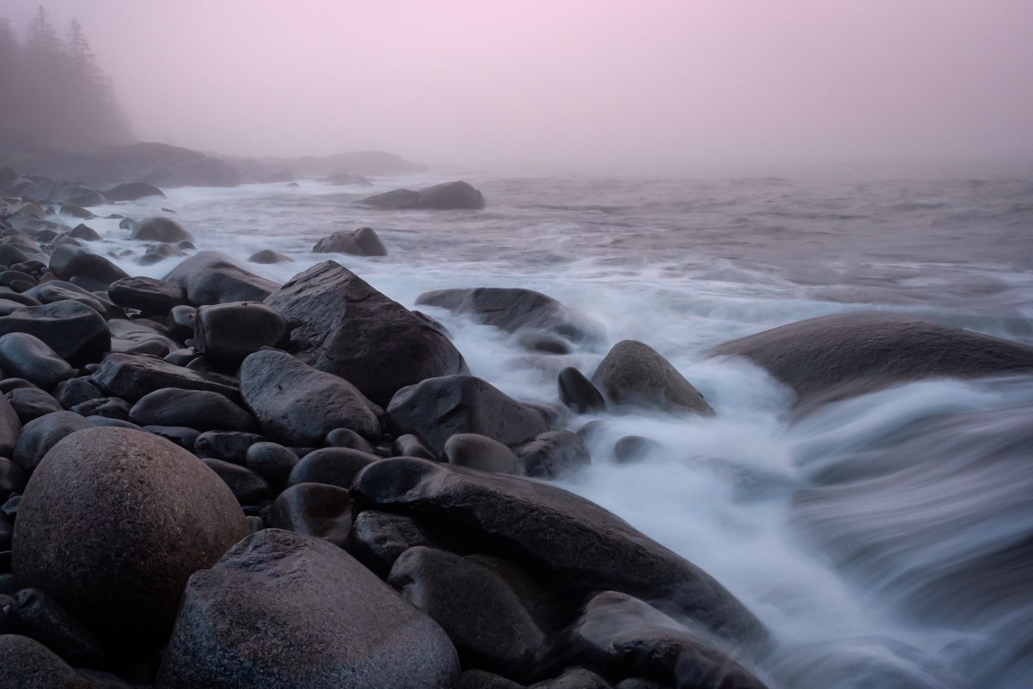 Misty coastal scene with waves crashing over smooth rocks in the morning fog at sunrise.