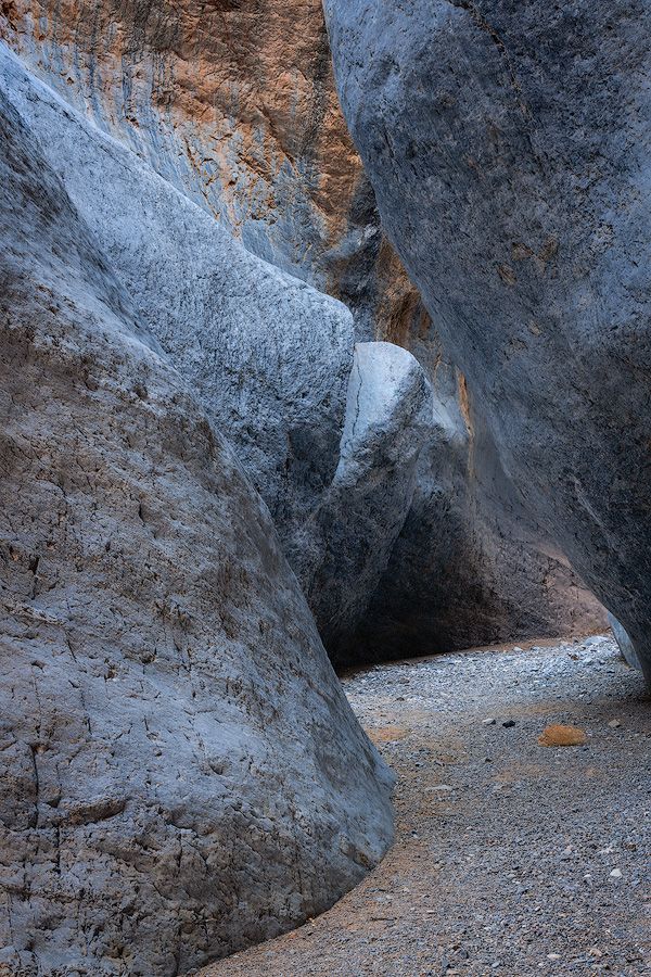 A narrow rocky canyon path winds through steep limestone walls with gravel and debris scattered on the ground.