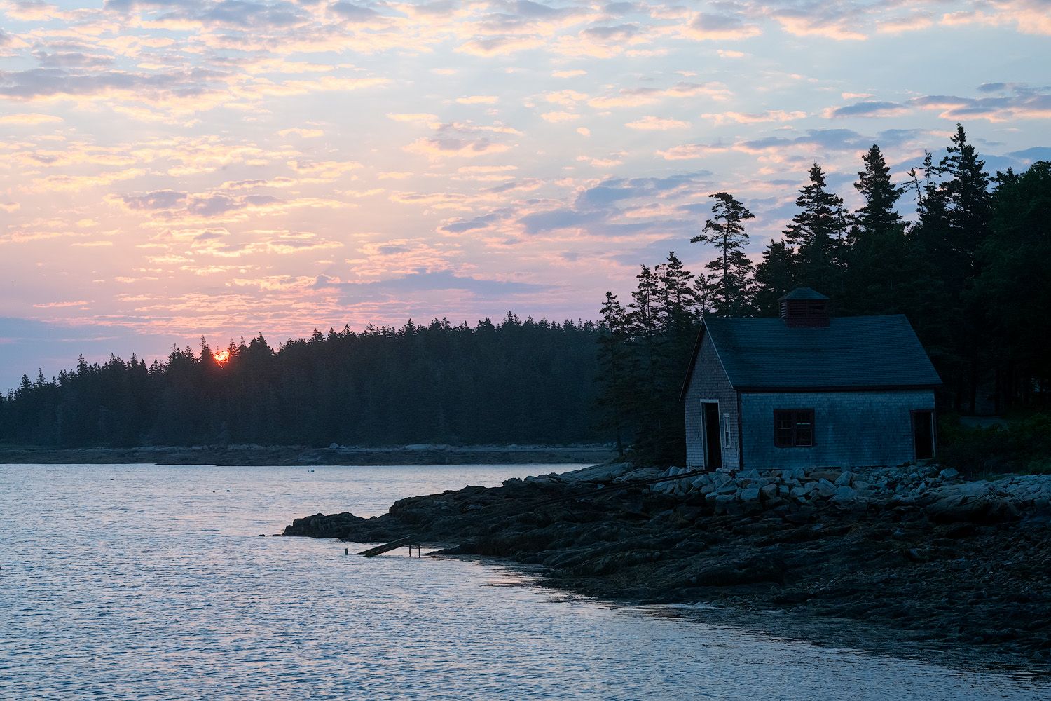 A small coastal cabin sits along a rocky shoreline at sunset with pine trees silhouetted against a pink and purple sky.