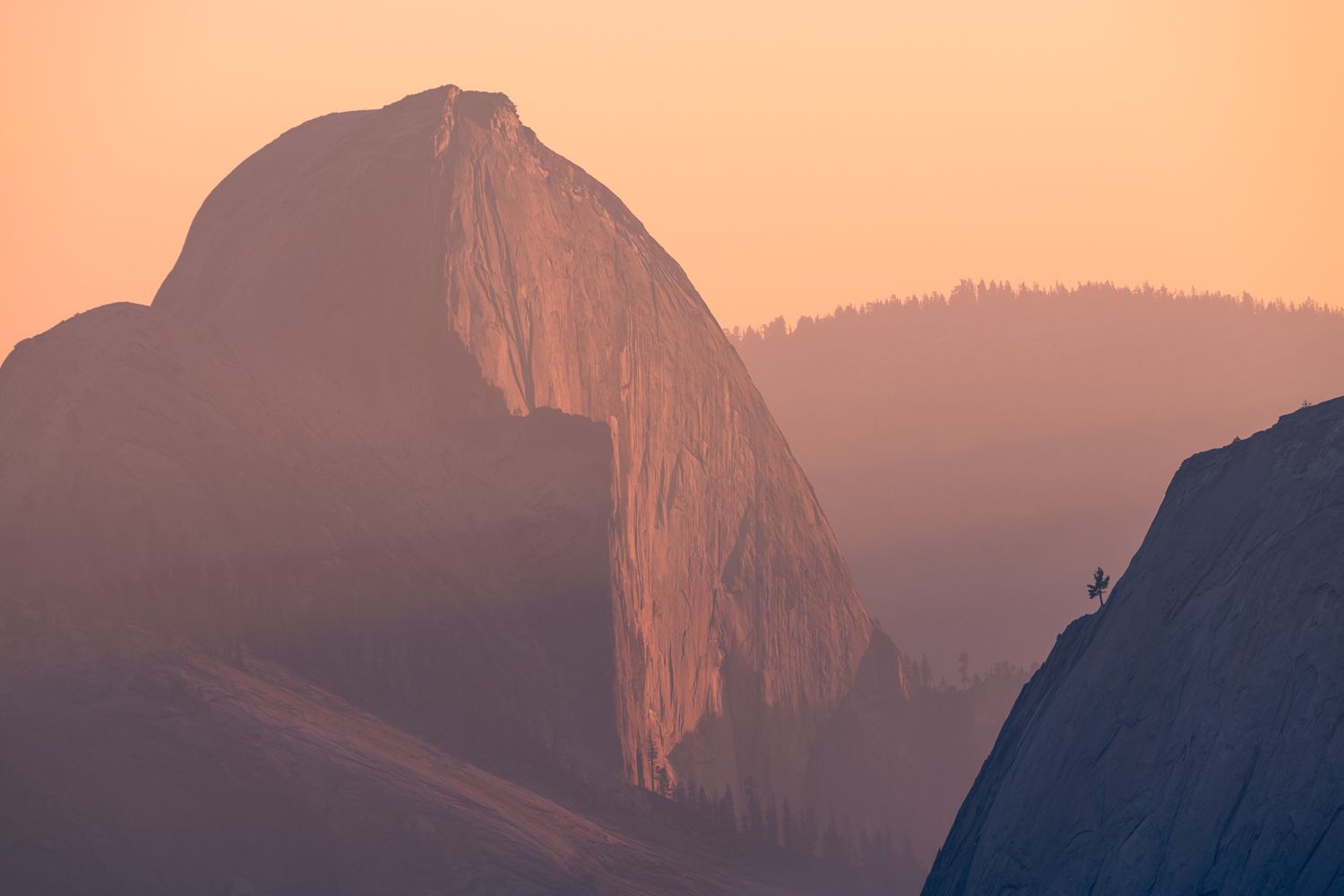 Half Dome's massive granite face glows orange in sunset light through smoky mountain air.