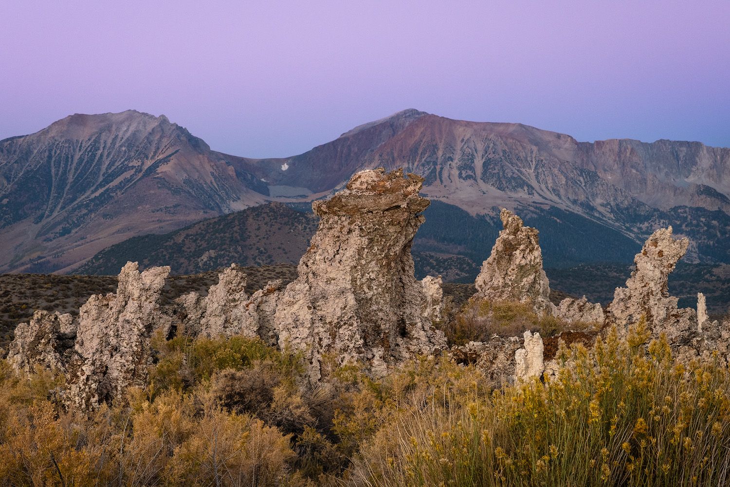 Dramatic rock formations rise against a purple-hued mountain landscape at sunrise with tufa formations.