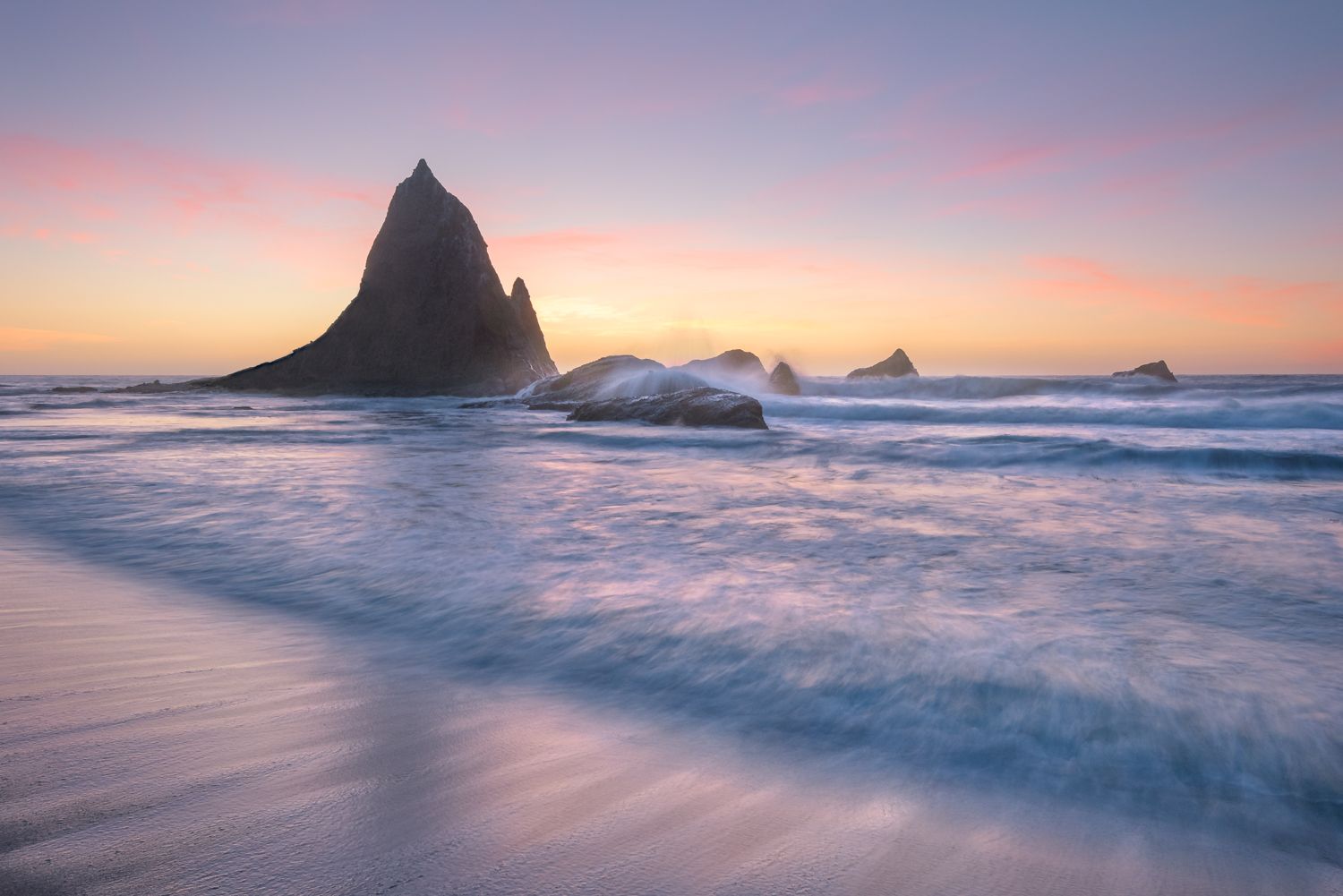 Dramatic coastal rock formations silhouetted against pastel sunset sky and rolling ocean waves.