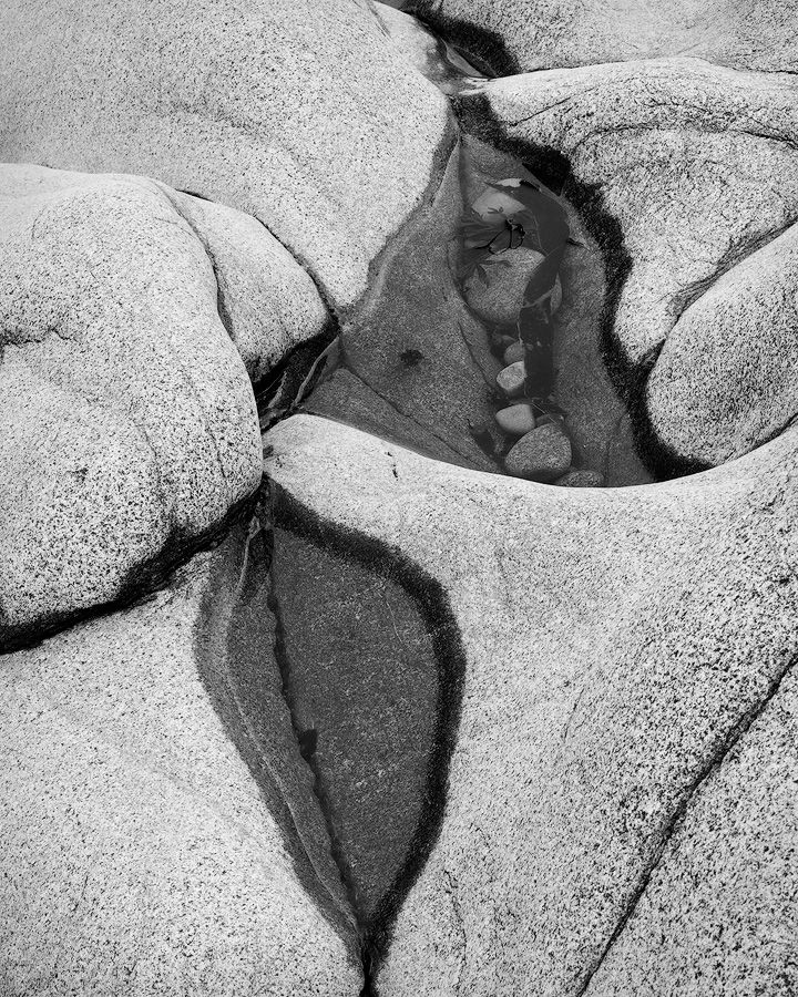 Black and white abstract photograph of water pooled between smooth granite rocks forming organic shapes and patterns.