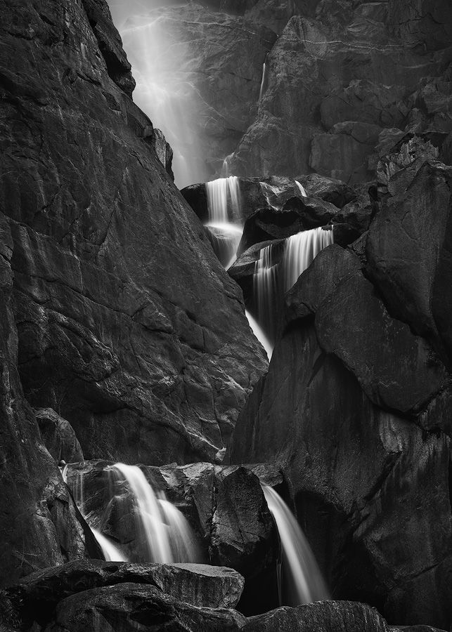 Long exposure black and white photograph of a waterfall cascading through rocky canyon walls in dramatic lighting.