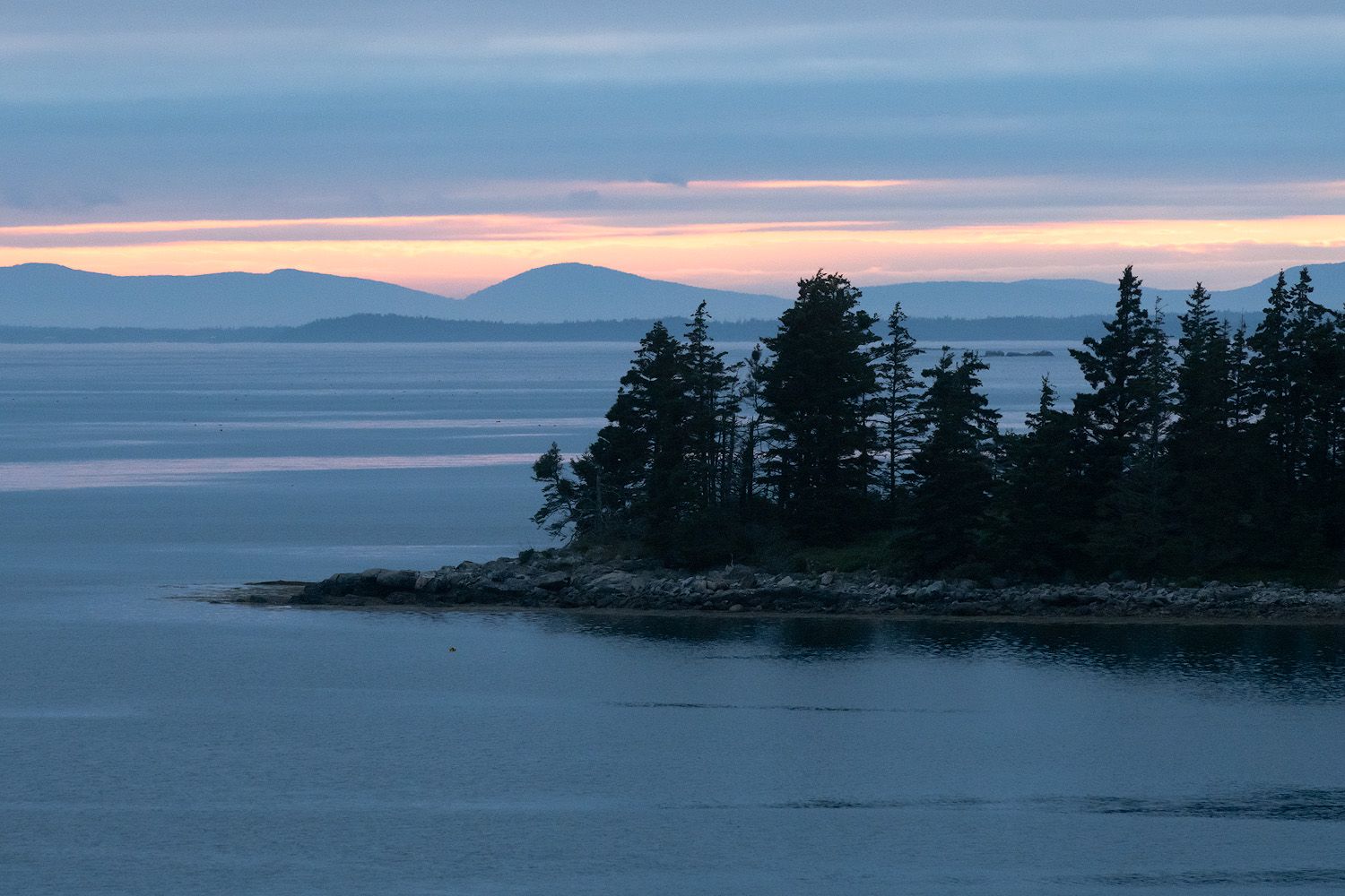 A silhouetted rocky coastline with evergreen trees against a tranquil dusk sky and calm ocean waters in Maine.