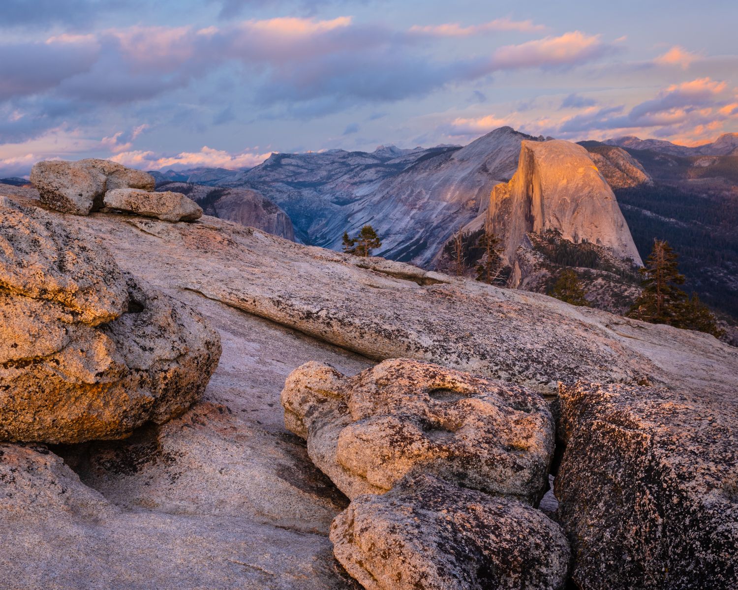 Granite rocks catch sunset light with Half Dome visible in Yosemite National Park's mountainous landscape.