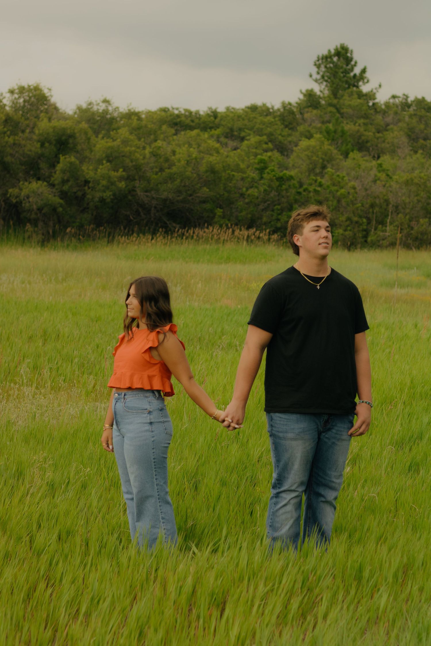 A couple holds hands while walking through a tall grassy field on a cloudy summer day.
