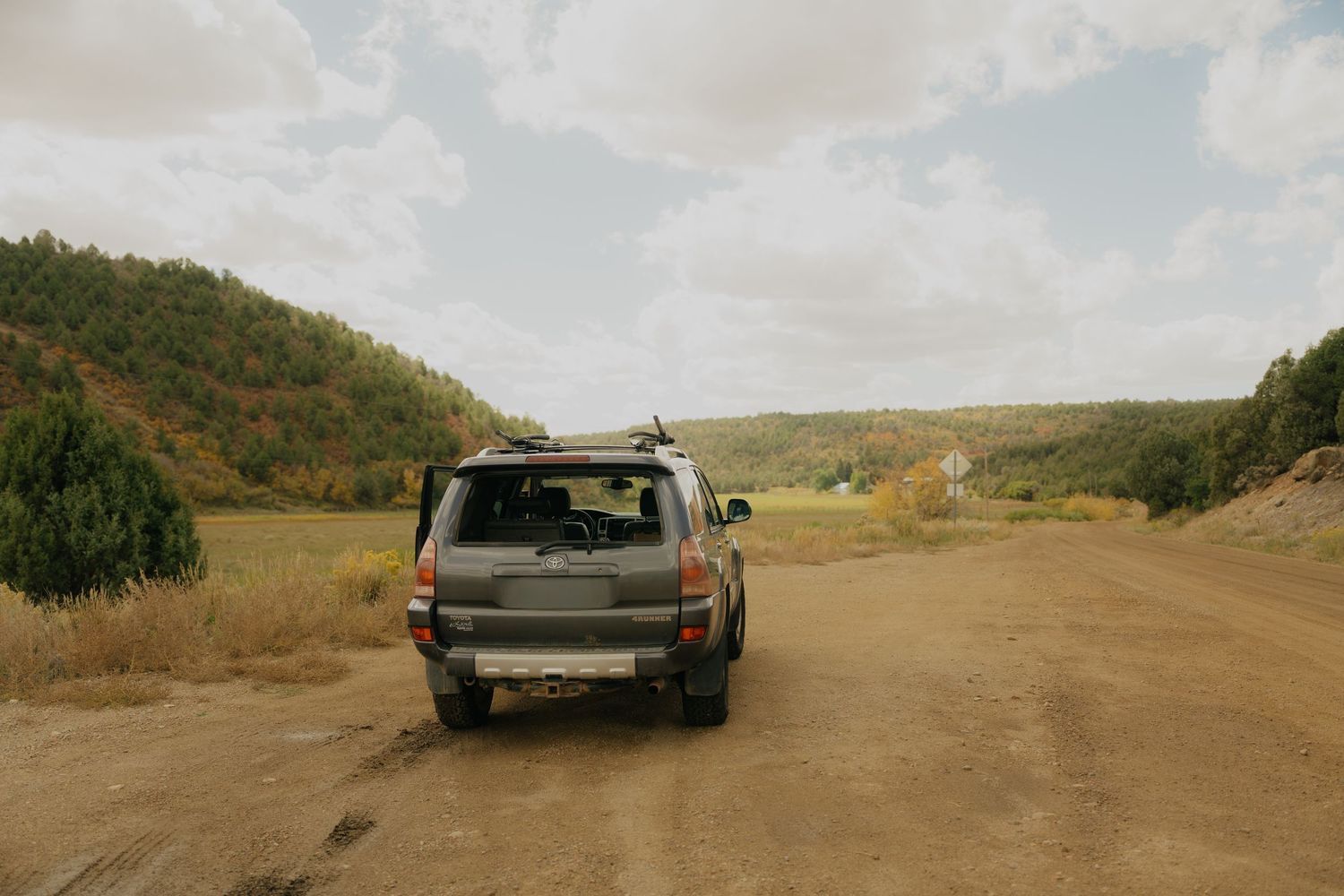 A vintage SUV parked on a dirt road surrounded by rolling hills and autumn foliage on a cloudy day.