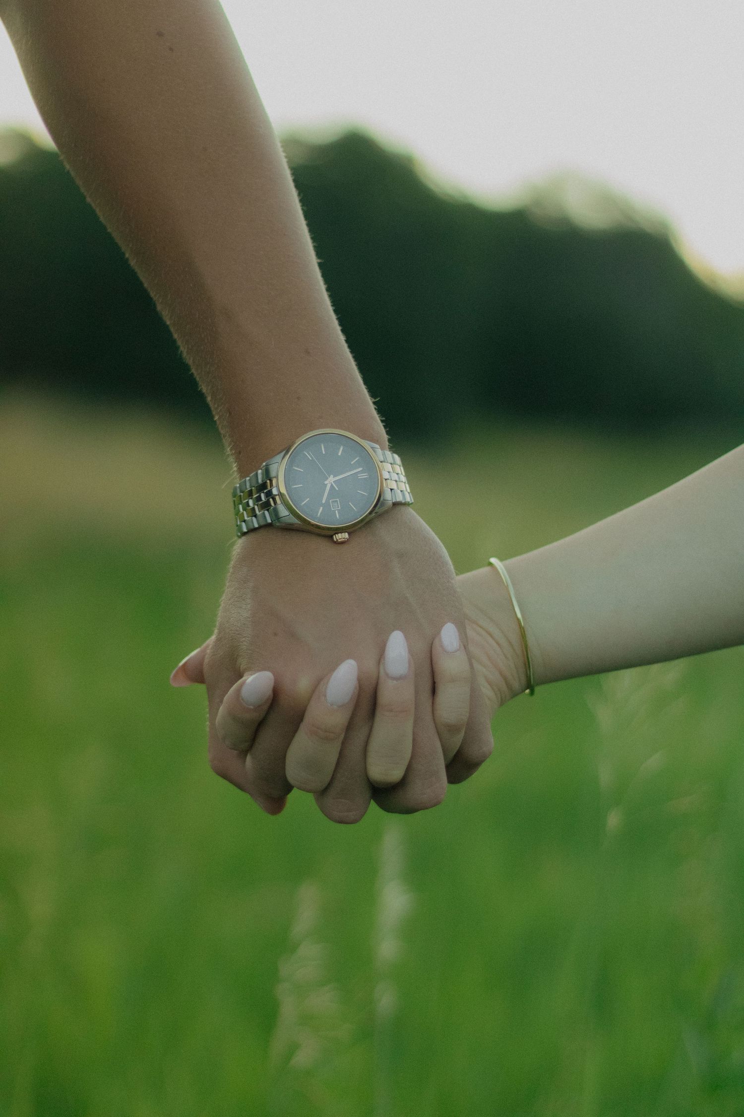 Close-up shot of two hands lovingly clasped together against a blurred green outdoor background, one wearing a silver watch.