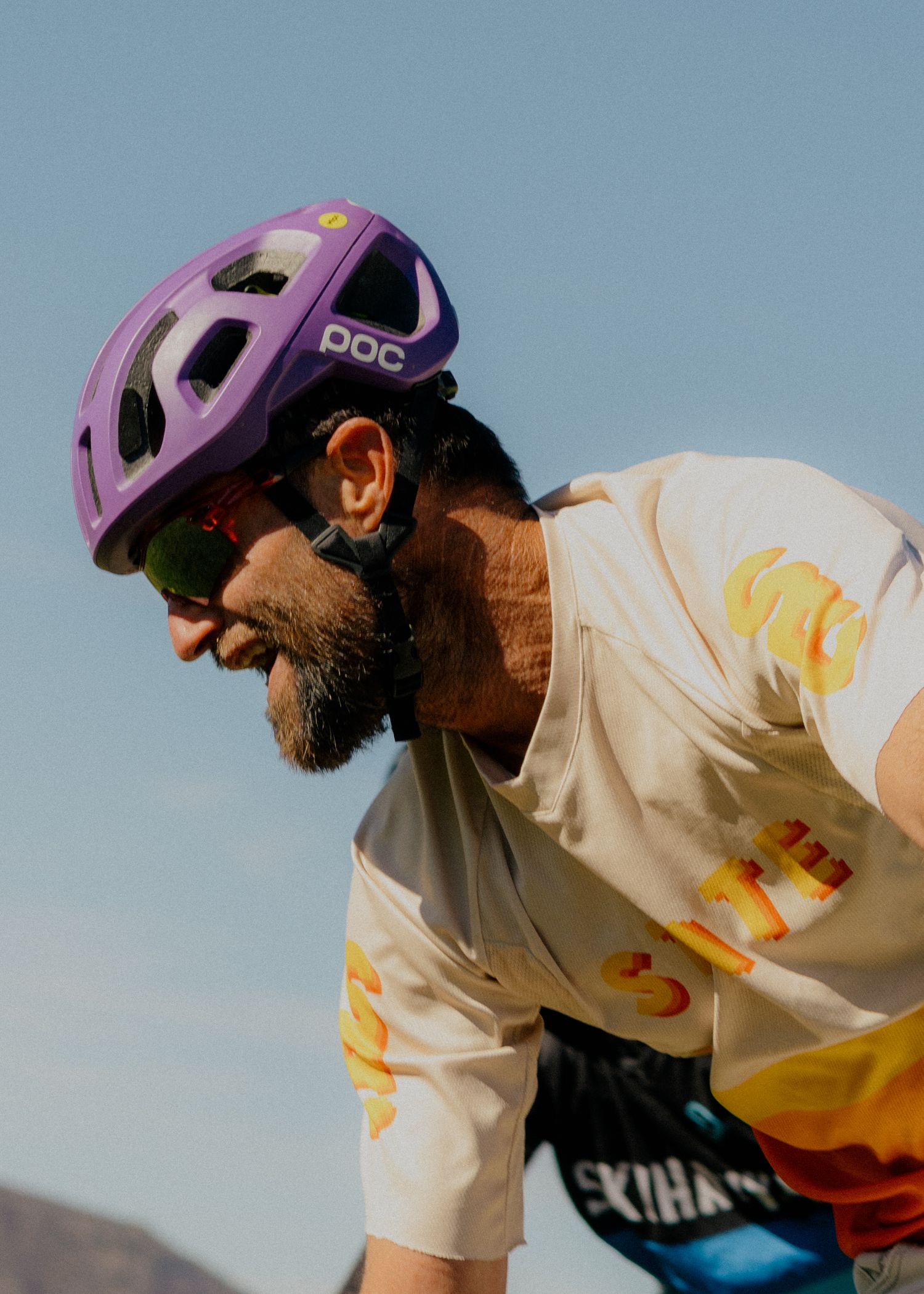 A cyclist in a purple helmet and white jersey rides against a blue sky backdrop.