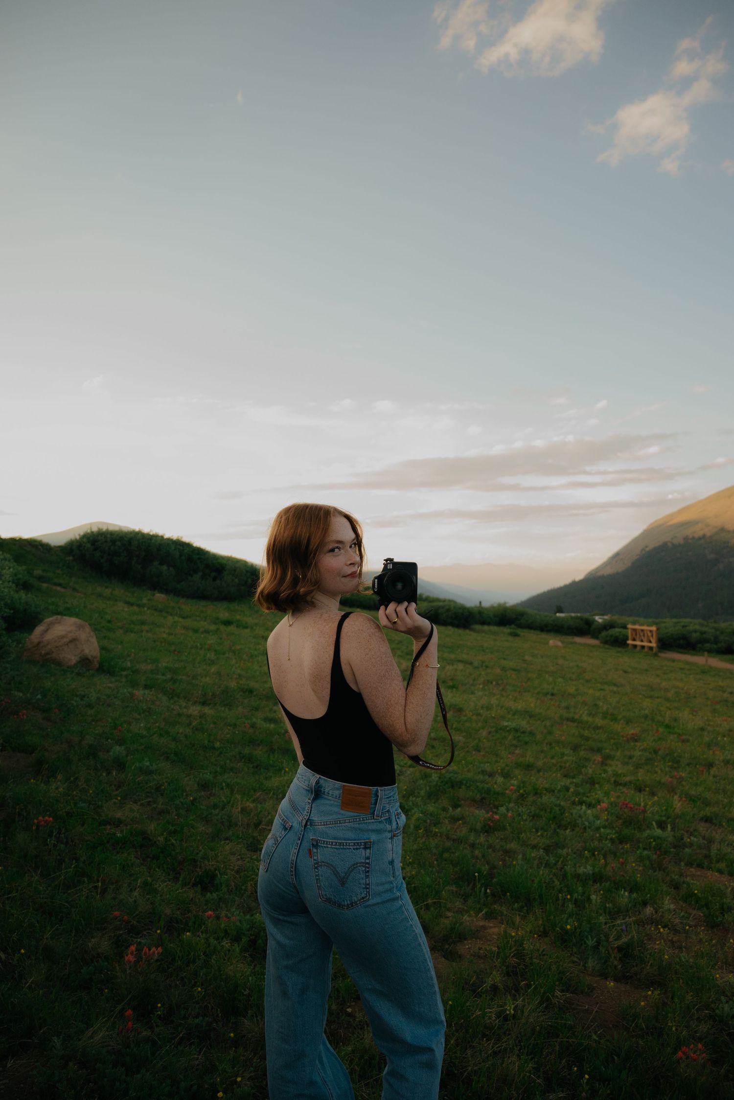 A photographer in denim jeans takes pictures in a scenic mountain meadow at dusk.