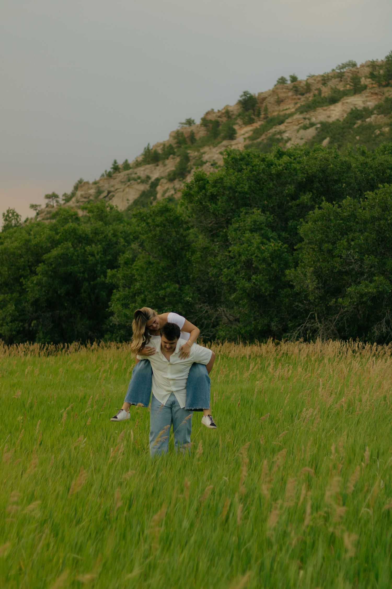 Two people in casual clothing playfully run through a lush green meadow with rolling hills in the background.