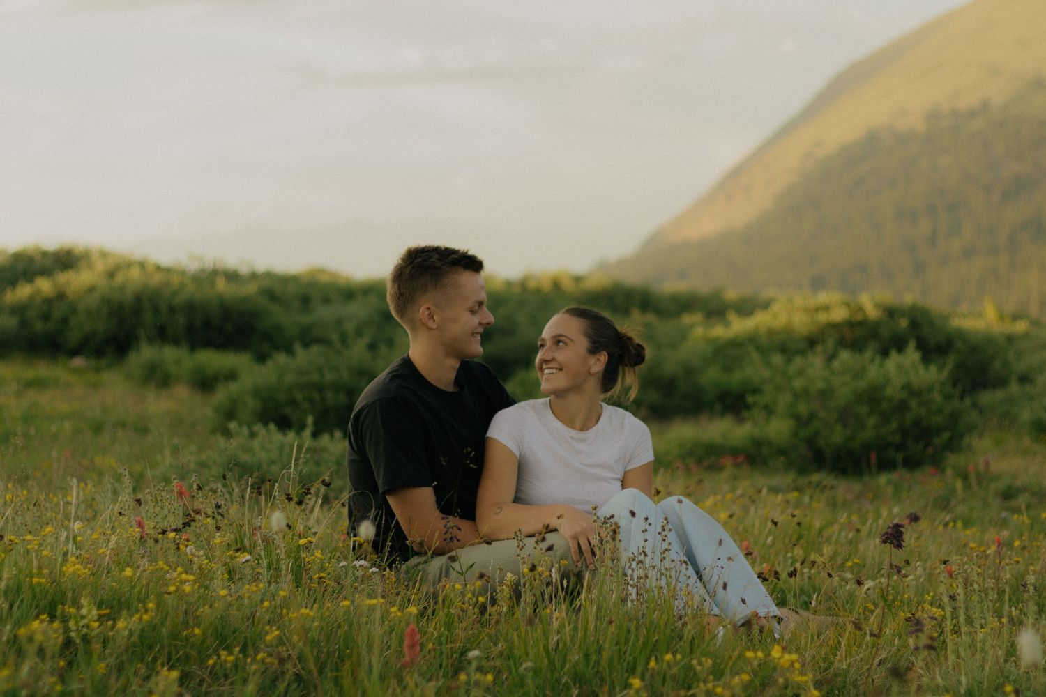 A couple sits together in a wildflower meadow at sunset with mountains in background.