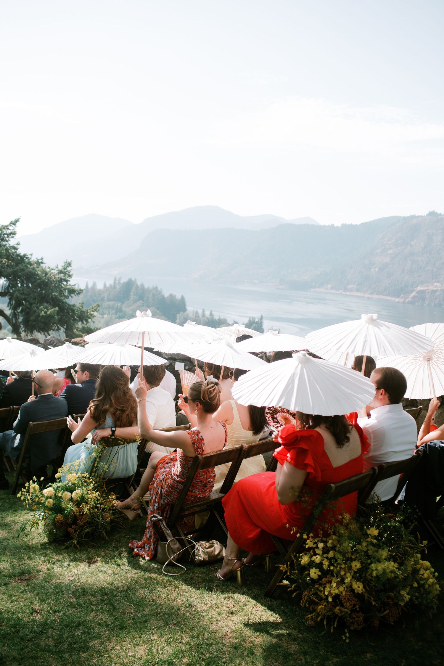 Wedding guests hold white parasols while sitting on a grassy hillside overlooking misty mountains during an outdoor ceremony in Hood River.