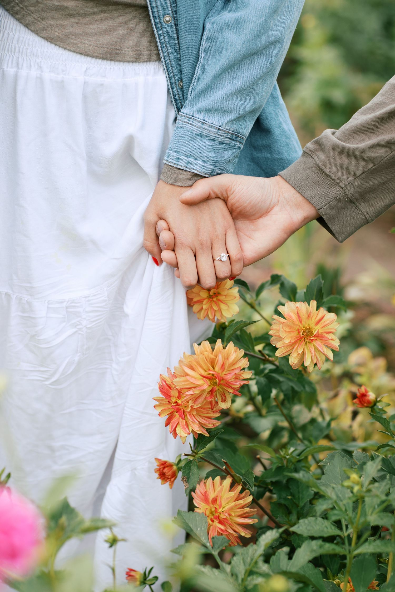 Two people holding hands tenderly next to blooming orange dahlias in a garden setting.
