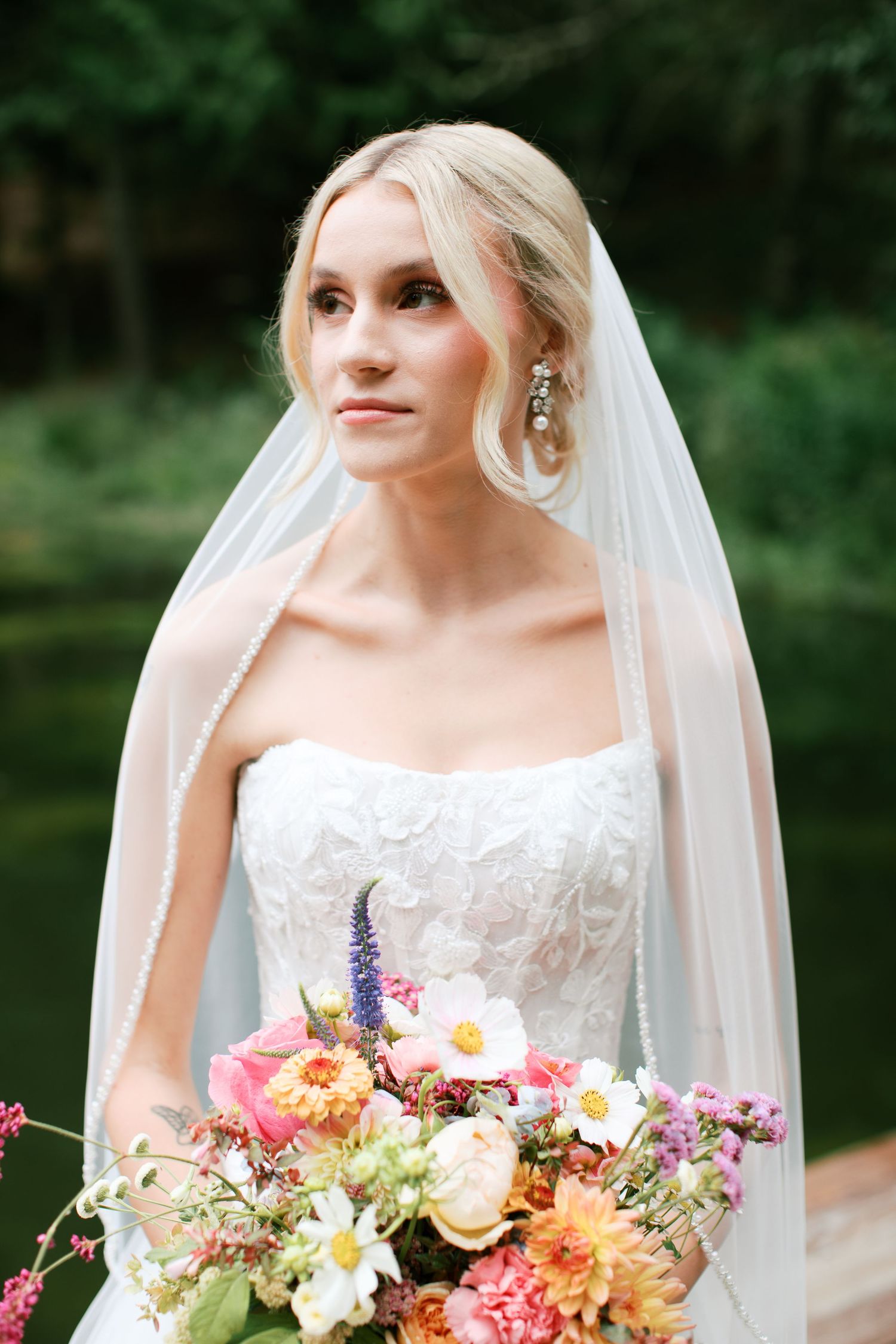 A bride in a white lace wedding dress holds a vibrant bouquet of pink, orange and purple wildflowers outdoors.