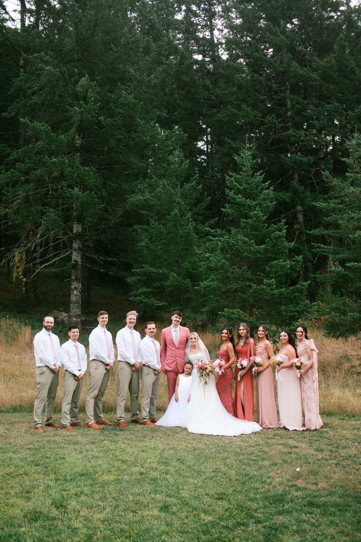 Oregon wedding party in coral and khaki attire poses together on grassy field with evergreen forest backdrop.
