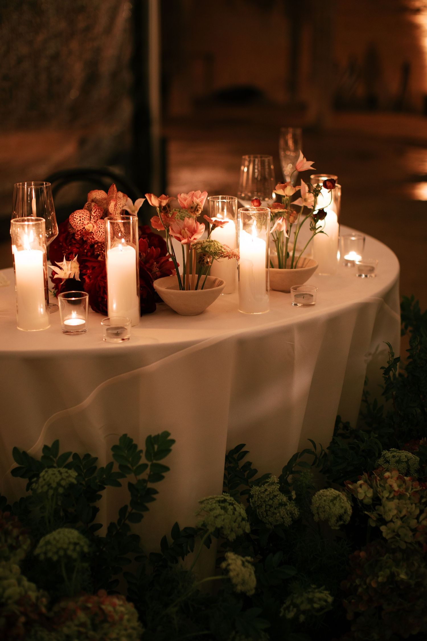 Romantic candlelit table setting with white linens and greenery decorations creates an intimate dining atmosphere.