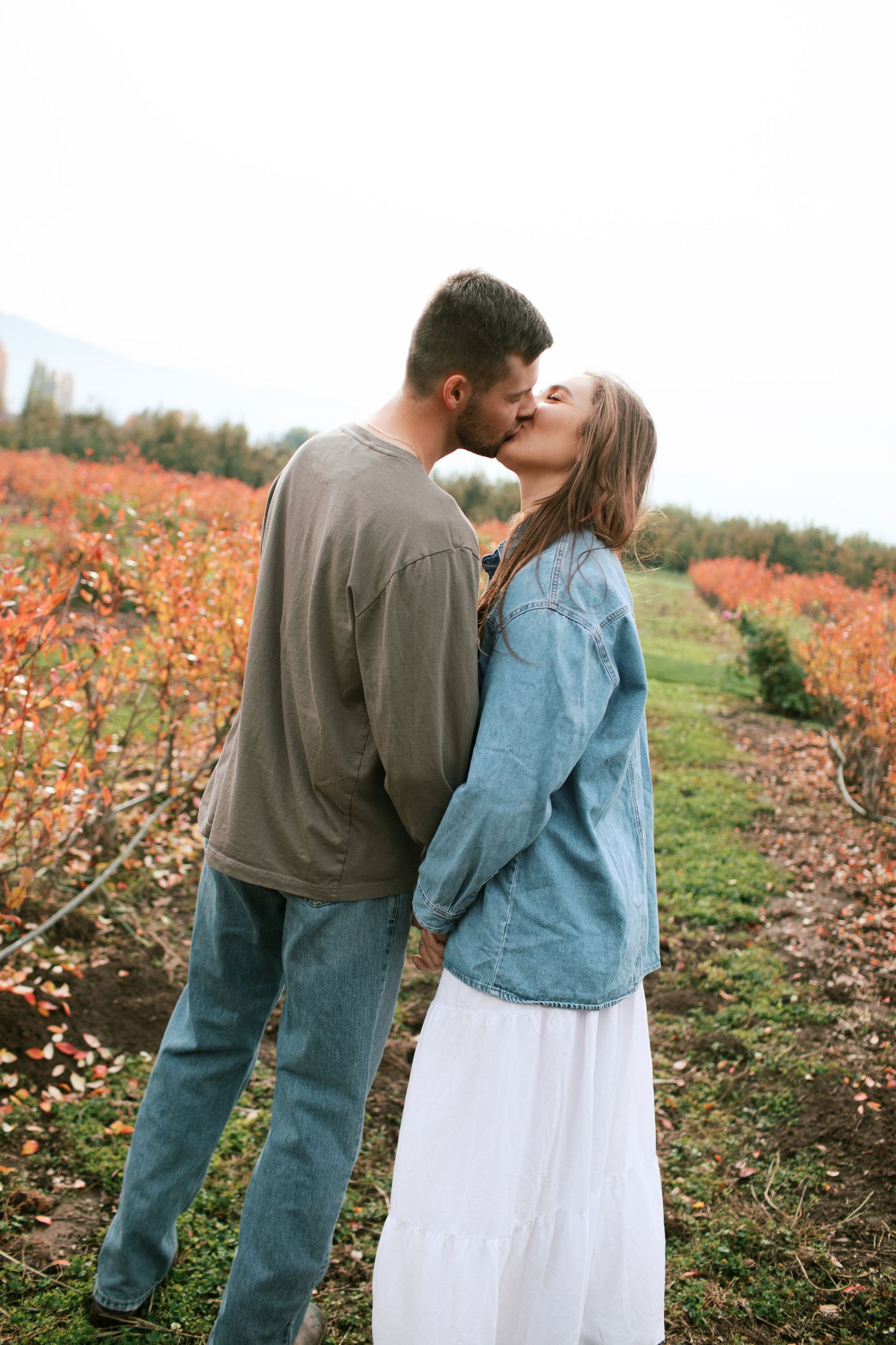 A couple shares a romantic kiss in a field with orange autumn foliage and trees in the background.