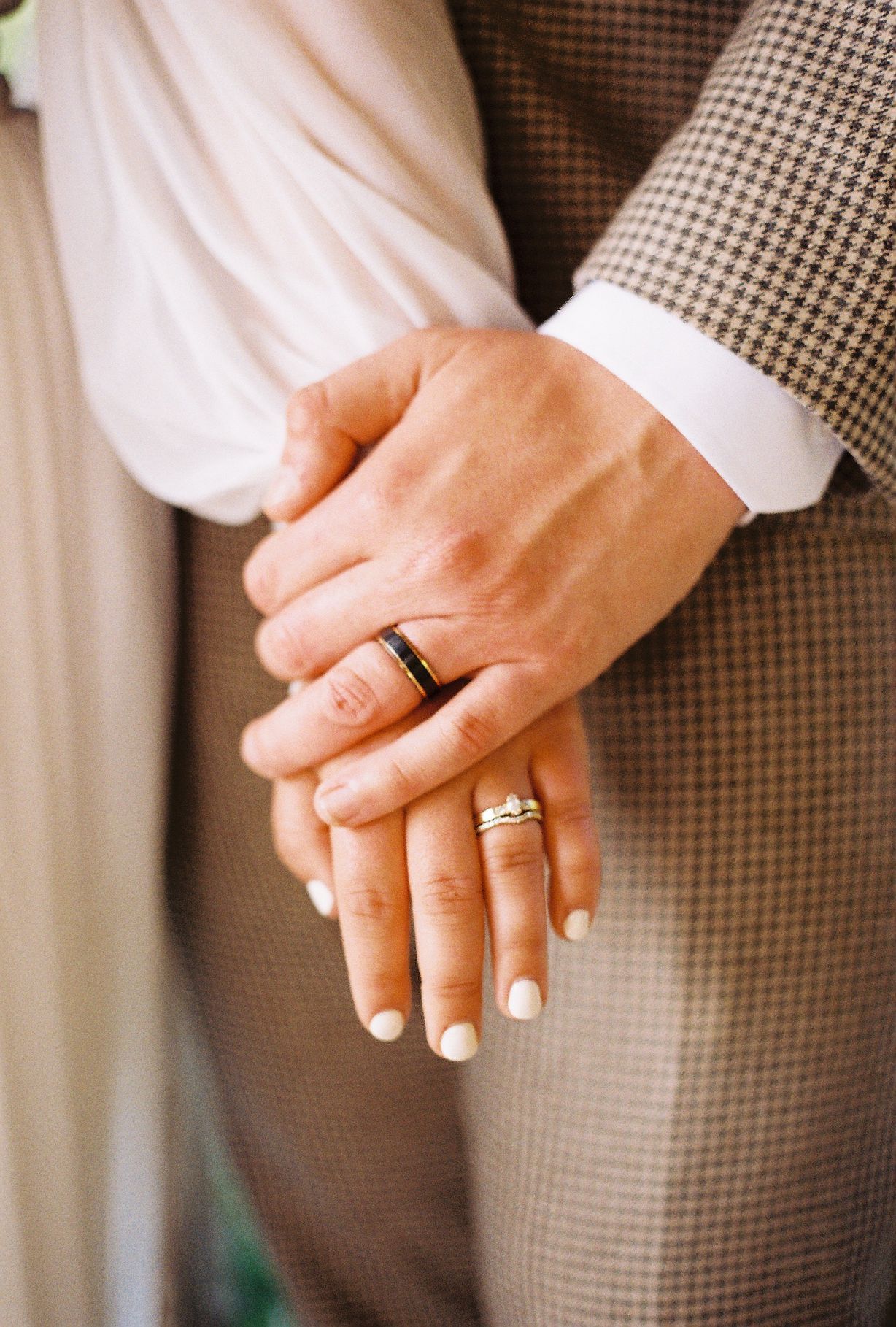 Close-up of newlywed couple holding hands, showcasing a black wedding band and gold stacked rings with white manicured nails.