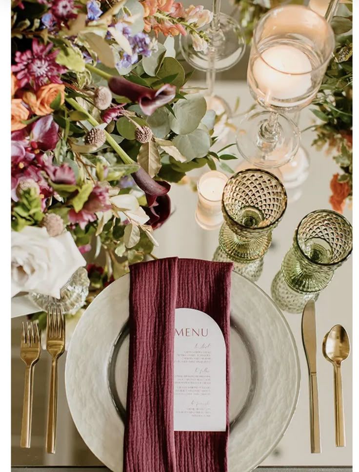 Elegant table setting with burgundy napkins, white plates, gold cutlery, floral arrangements and glowing candlelight in vintage glasses.