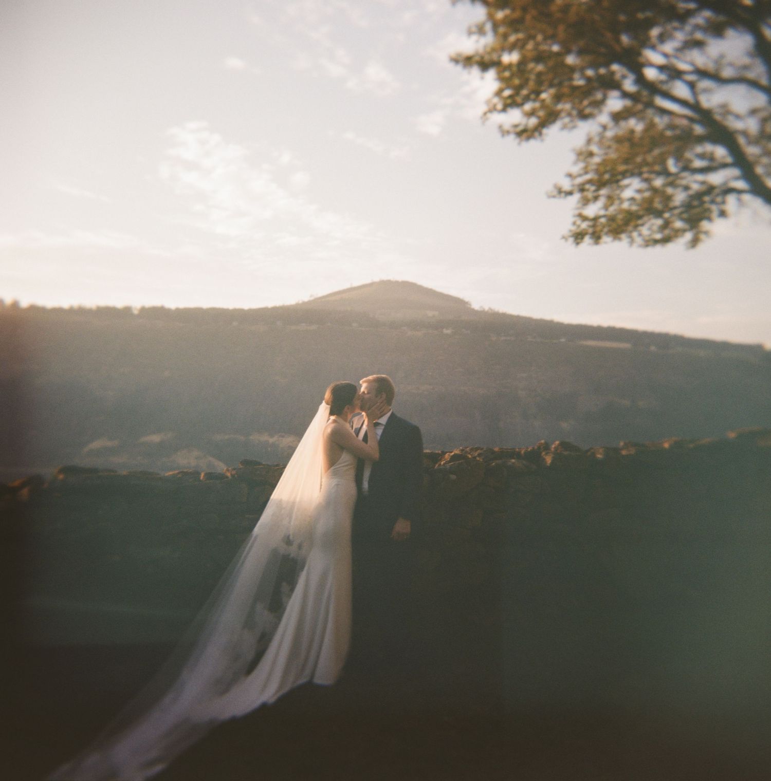 A couple in wedding attire stands silhouetted against a sunset mountain landscape in a romantic outdoor setting.