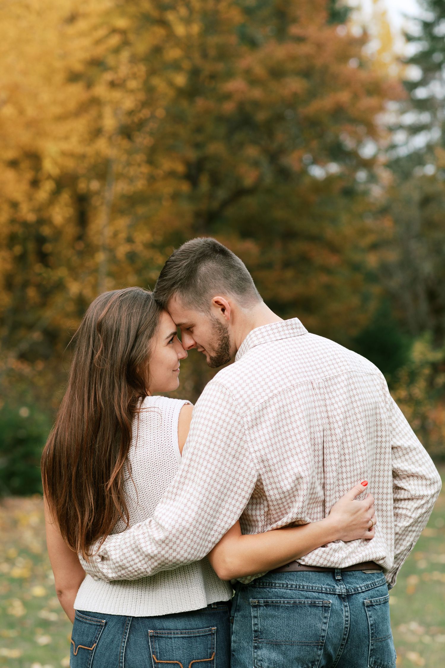 A couple embraces while standing in a park with golden autumn foliage in the background.