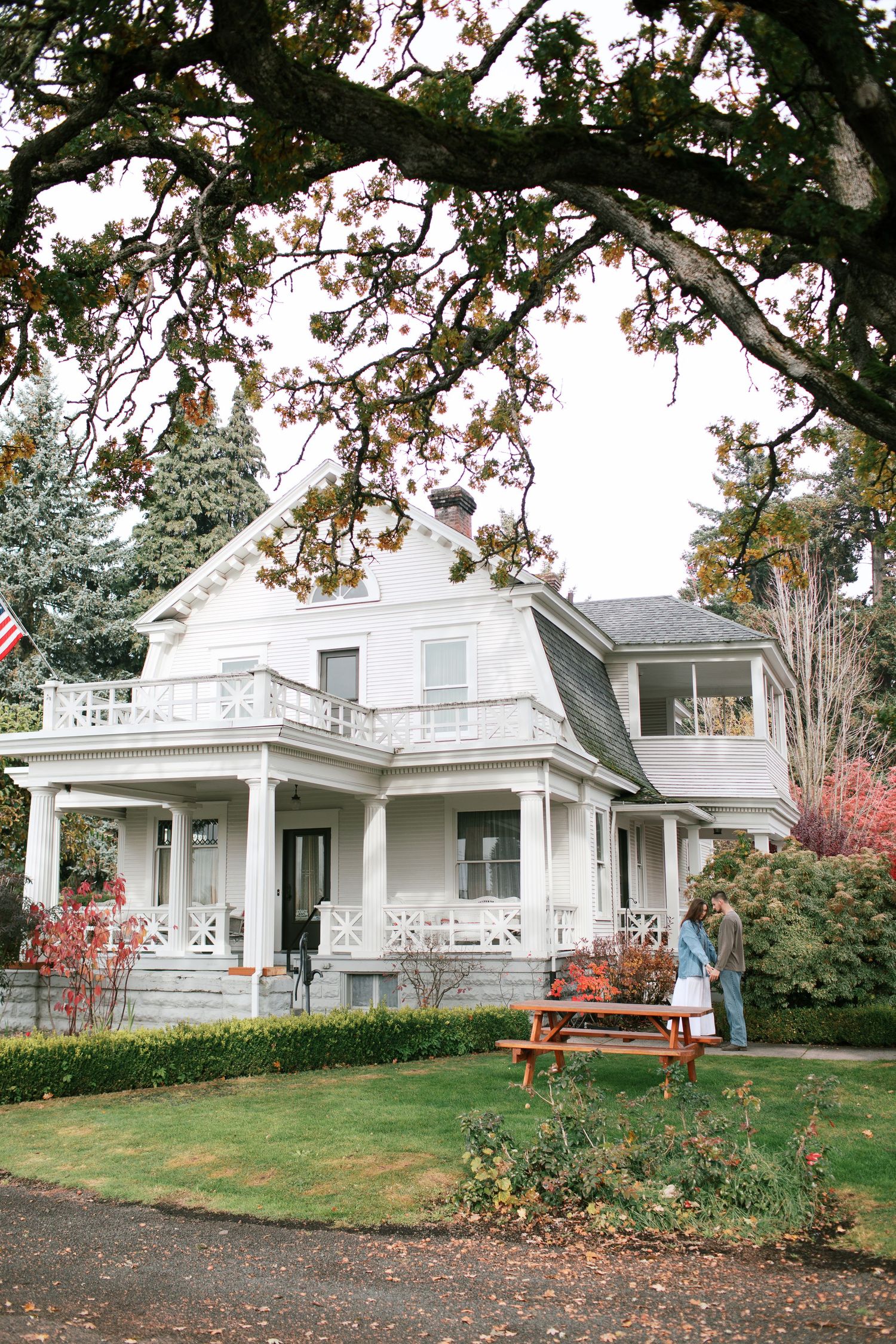 White historic Victorian mansion with wraparound porch and manicured lawn surrounded by autumn trees and picnic tables.