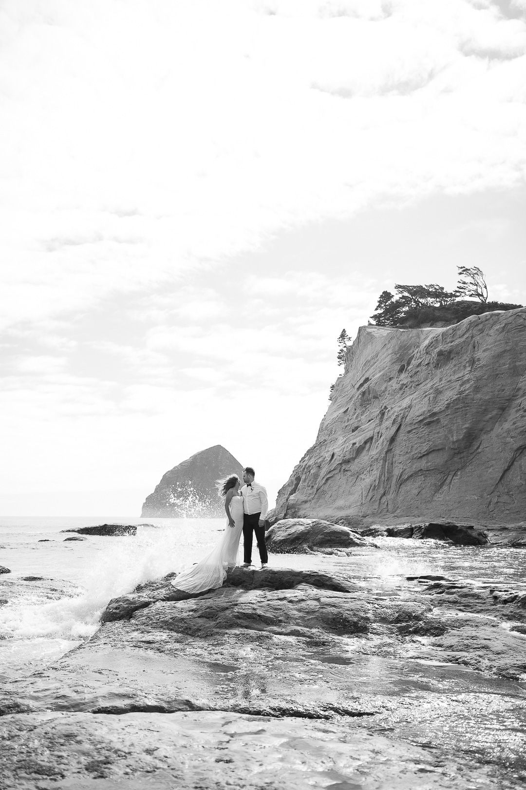 A couple embraces on rocky coastal cliffs with crashing waves and dramatic seaside landscape in black and white.