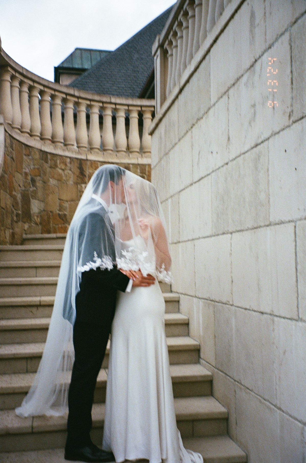 A romantic wedding couple shares an intimate moment on elegant stone stairs with a flowing veil creating a dreamy atmosphere.