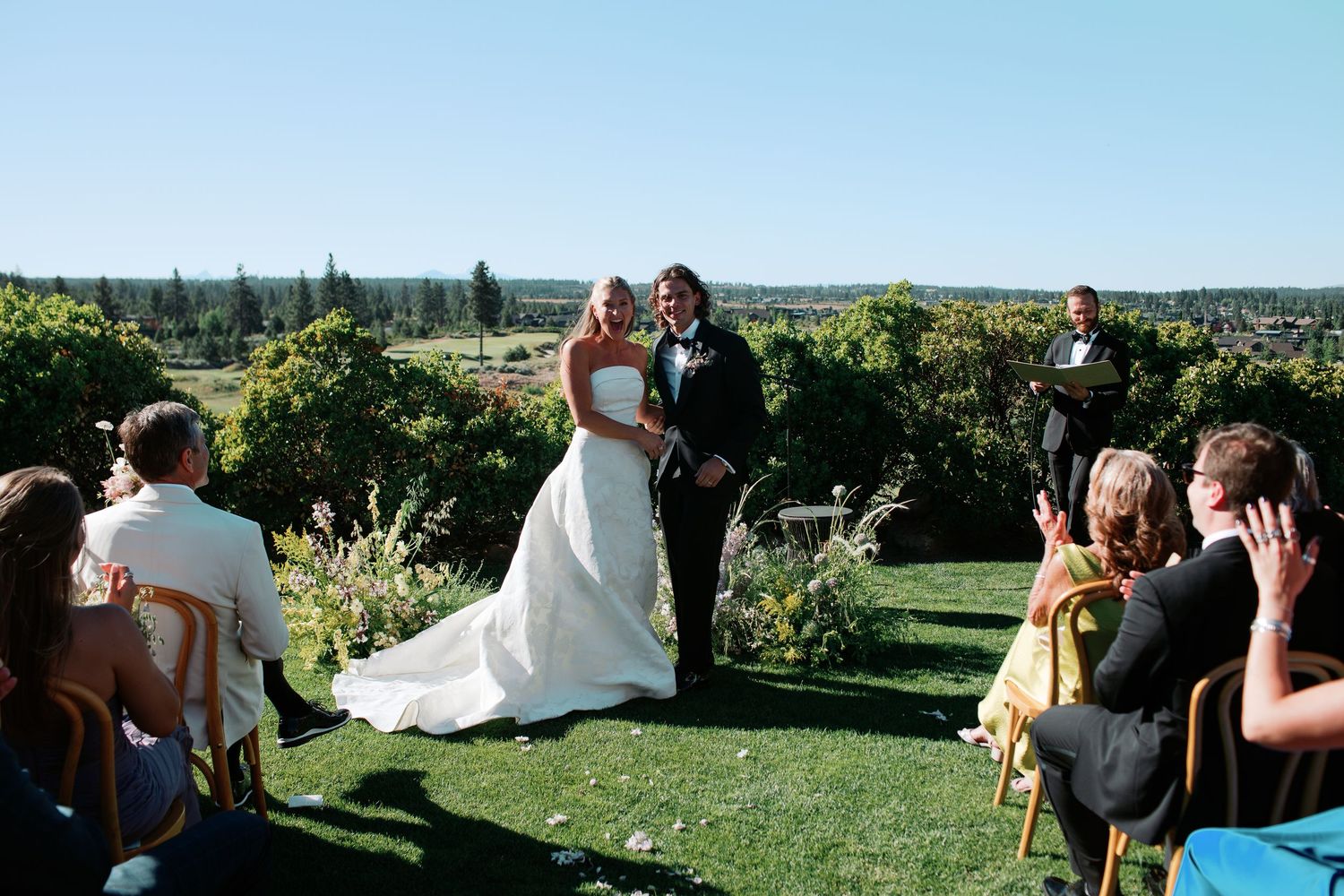 Bride and groom smile at outdoor wedding ceremony on lush green lawn with scenic vineyard views under clear blue sky.