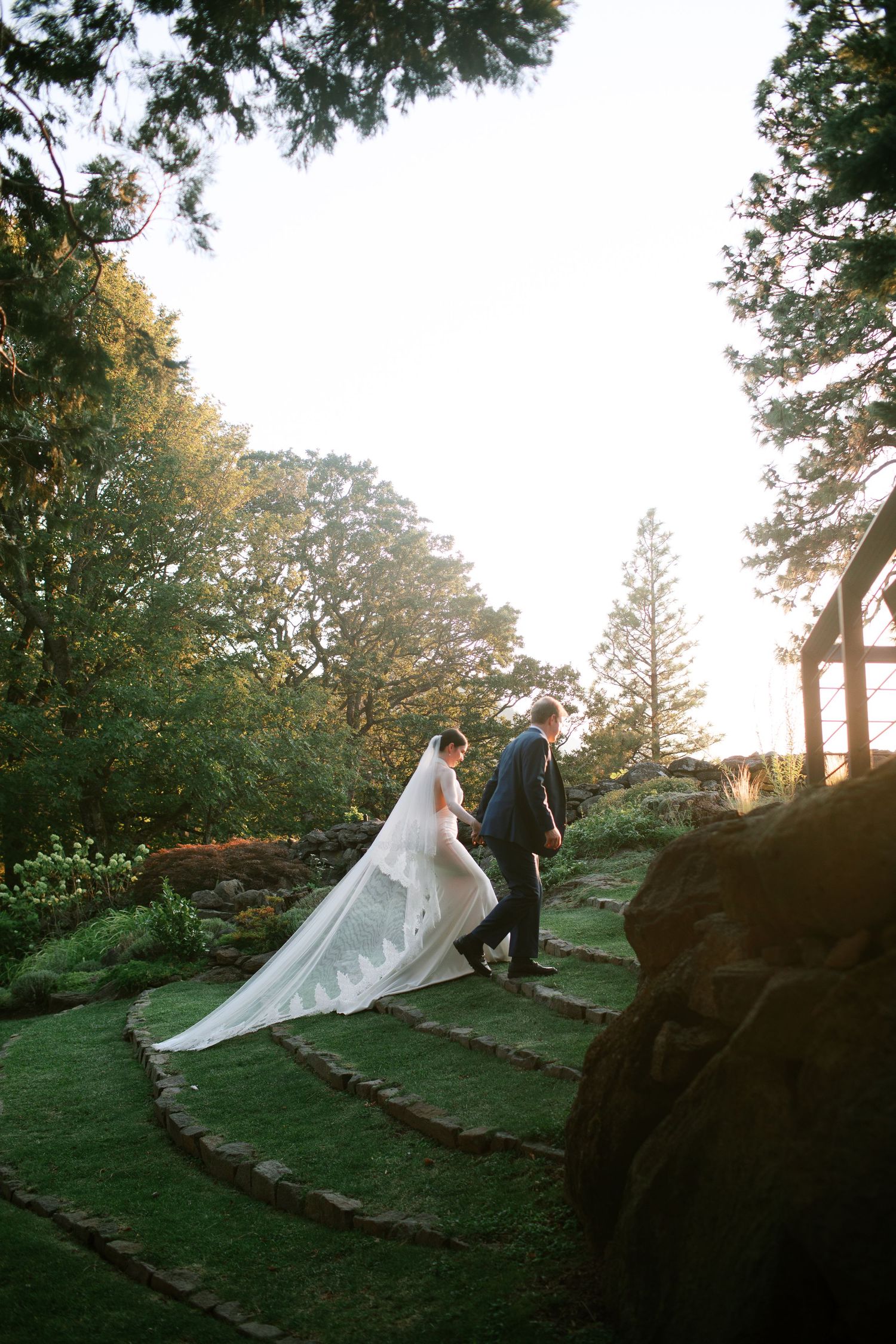 A flowing white wedding veil billows dramatically in the wind against a scenic hillside garden backdrop at sunset.