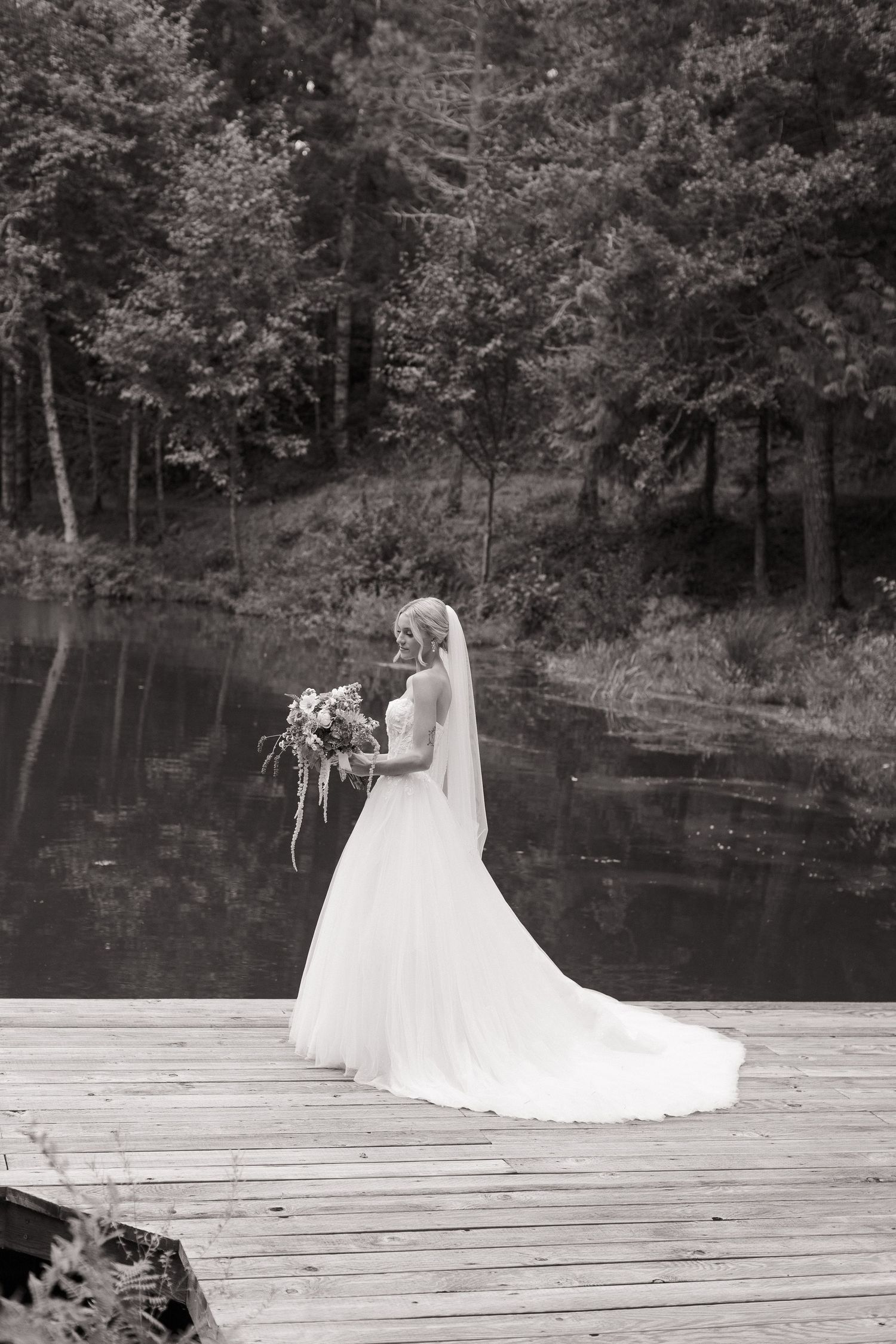 A bride in a flowing white wedding dress poses on a wooden dock by a serene lake surrounded by trees in black and white.