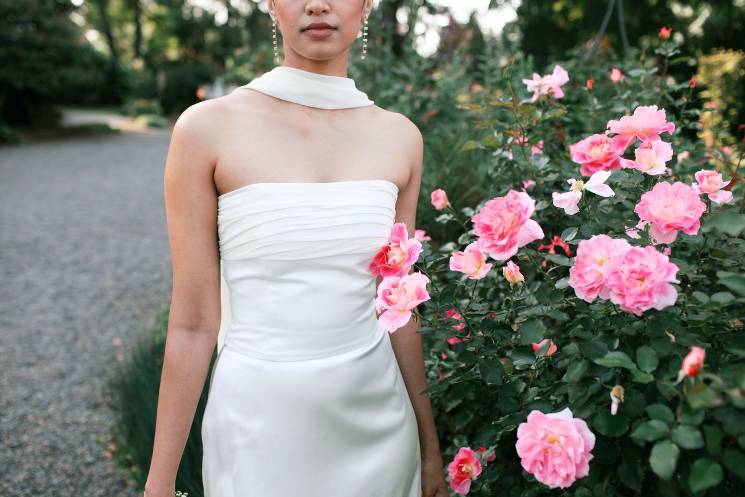 A bride in a white strapless wedding dress stands among blooming pink roses in a garden setting.